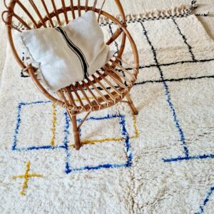 Ce tapis Beni Ouarain coloré est entièrement réalisé en laine naturelle noué à la main de brins de laine naturelle de coloris blanc-écru, jaune ocre, bleu et noir.