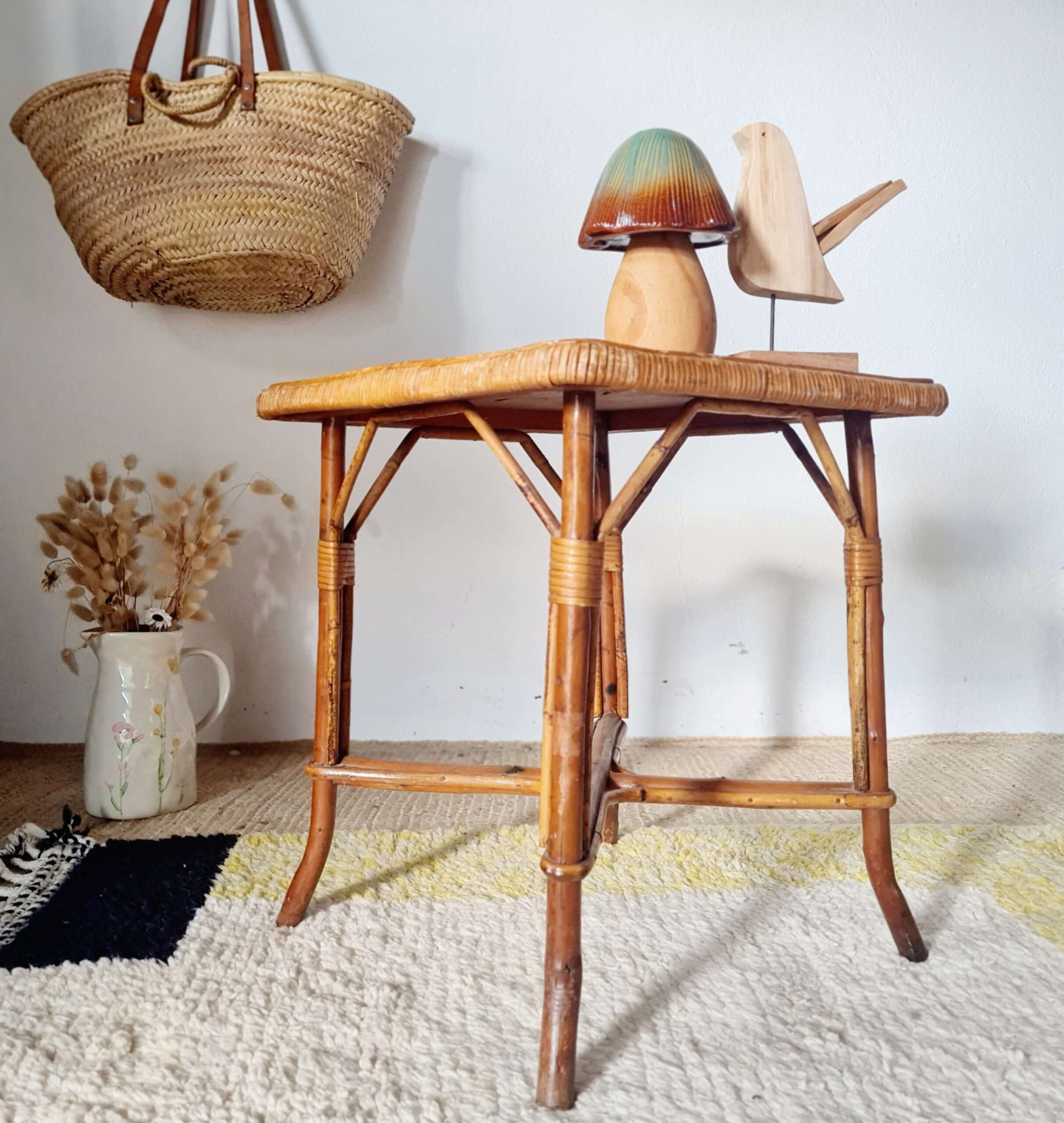 Vue globale de trois-quarts d'une table d'appoint en rotin style bohème, décorée d'objets en bois et entourée d'un pichet en grès et d'un couffin suspendu.
