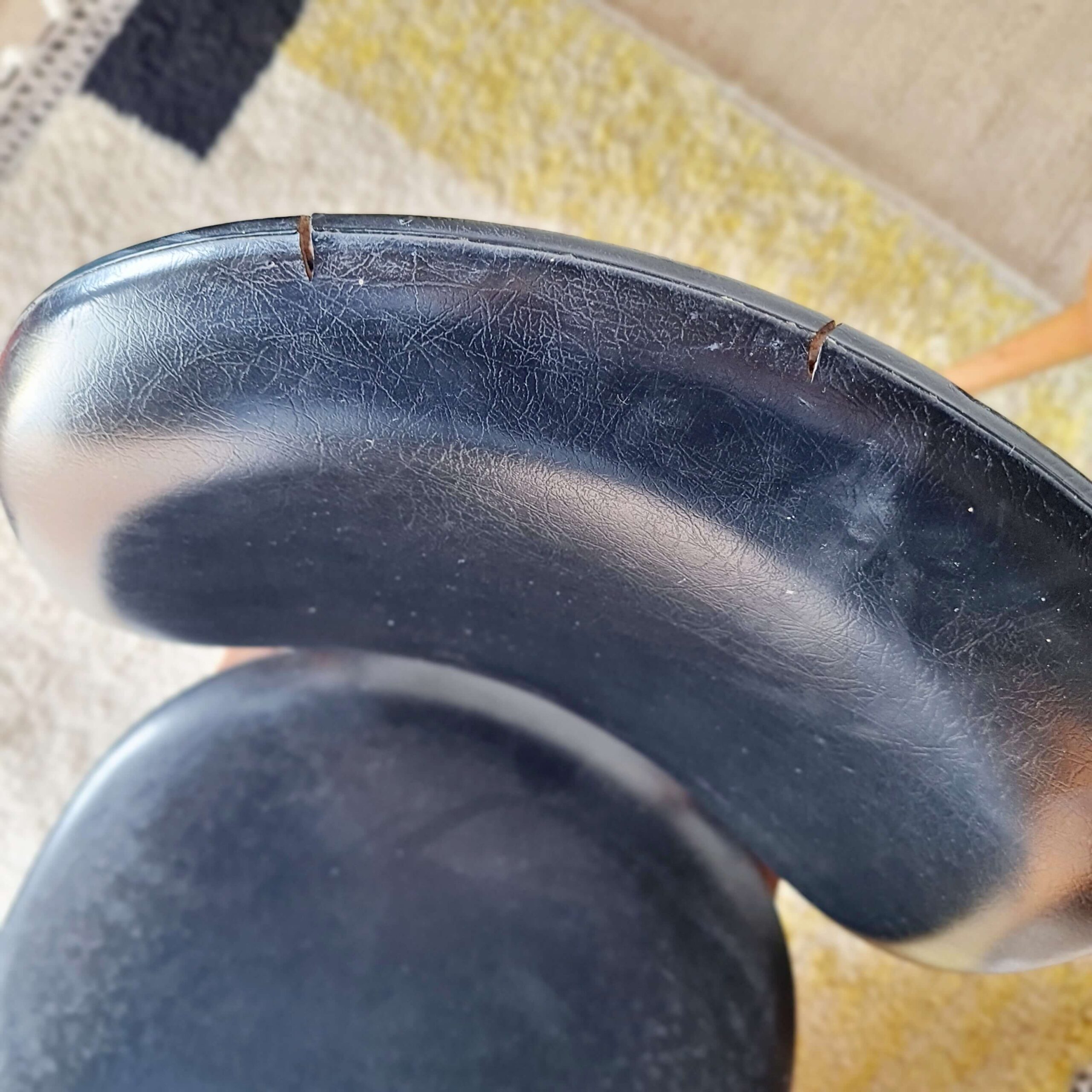 Gros plan détaillé sur le bord d'une assise de chaise vintage en simili-cuir noir, montrant deux petites coupures et de légères craquelures dues à l'usure du temps.