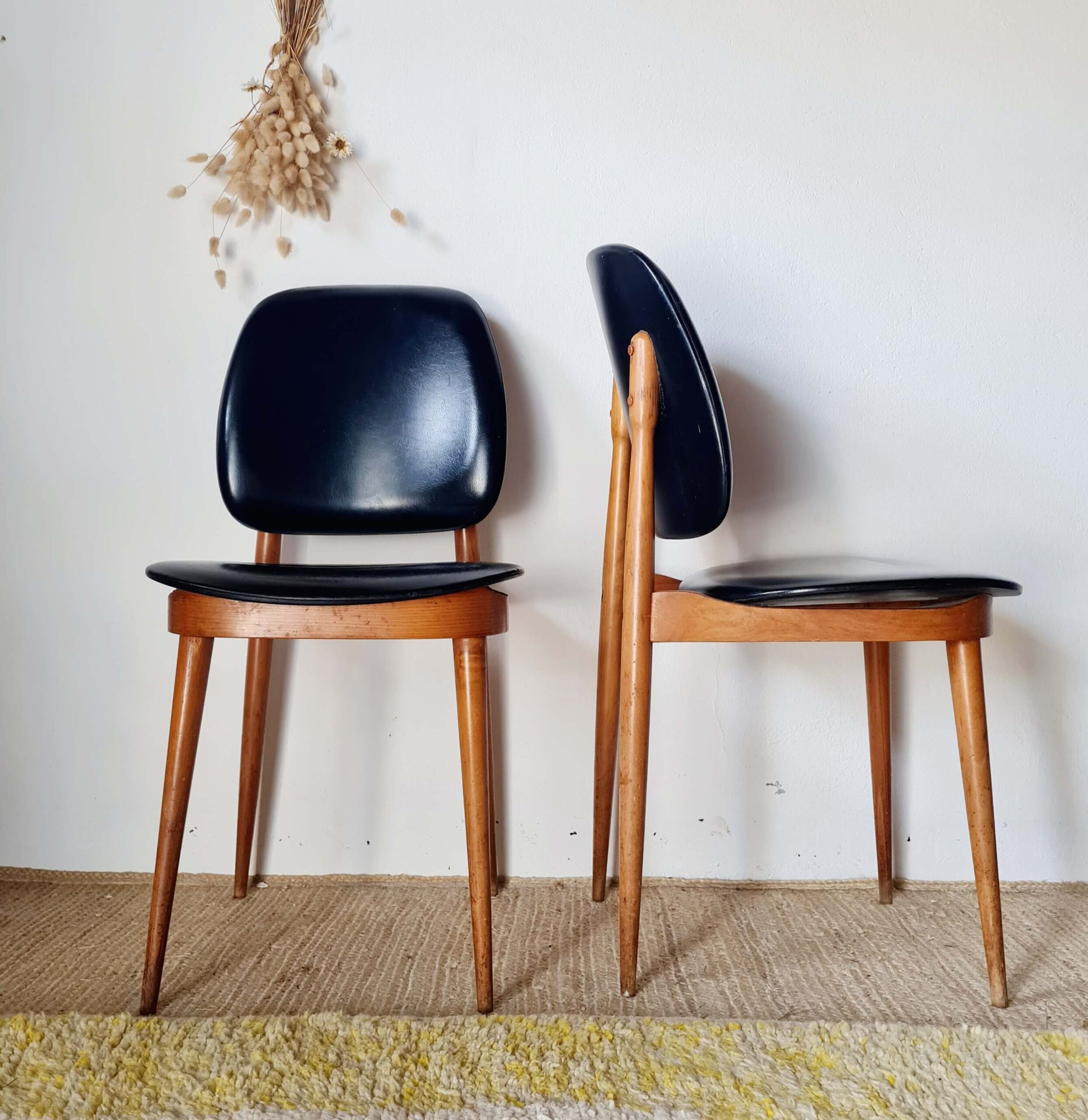 Paire de chaises rétro des années 1960 avec structure en bois et revêtement noir, l'une présentée de face et l'autre de profil devant un mur blanc.