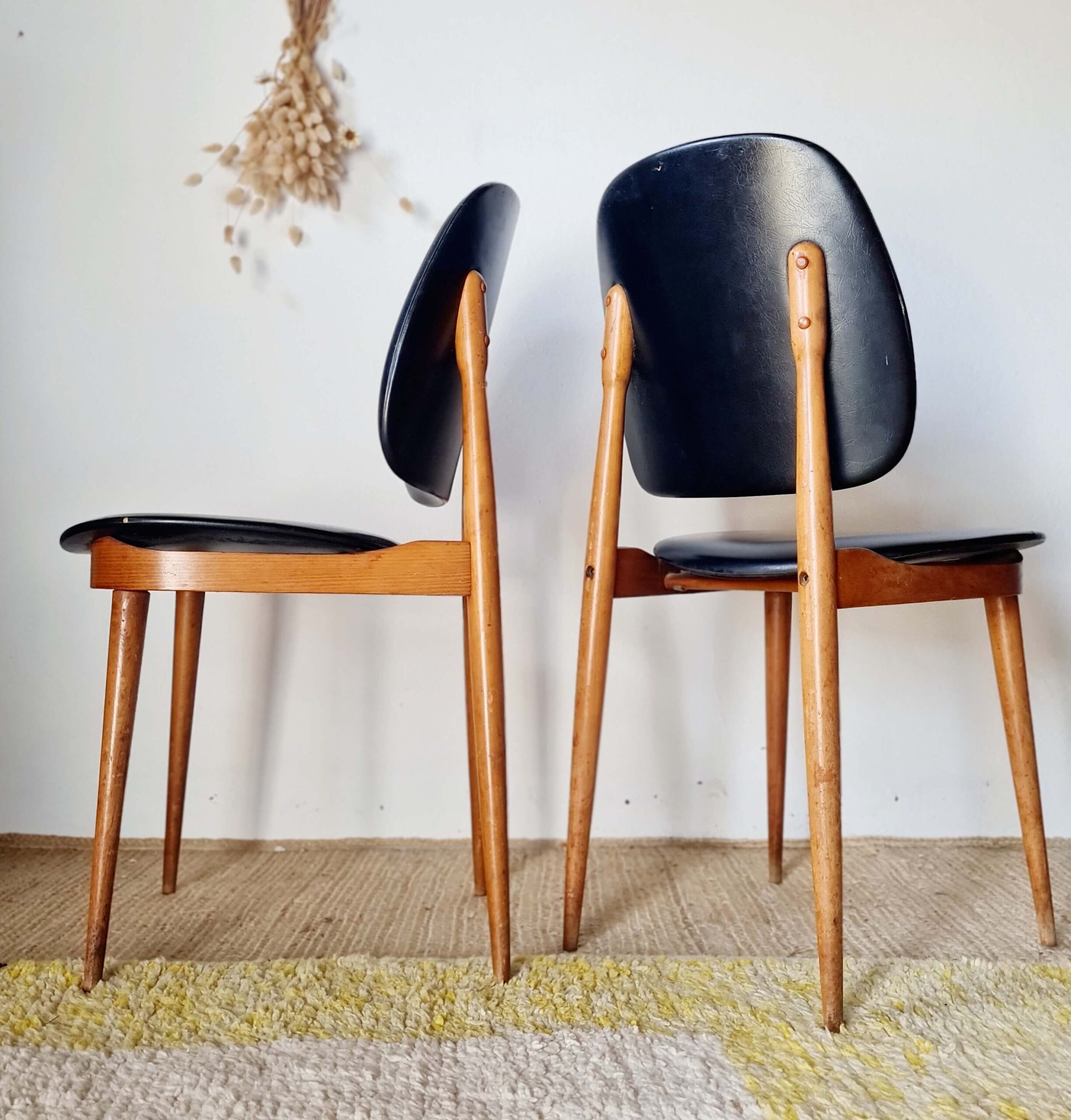 Paire de chaises de style moderniste des années 60, l'une de profil et l'autre de face, avec structure en bois clair et revêtement noir, posées sur un tapis texturé.