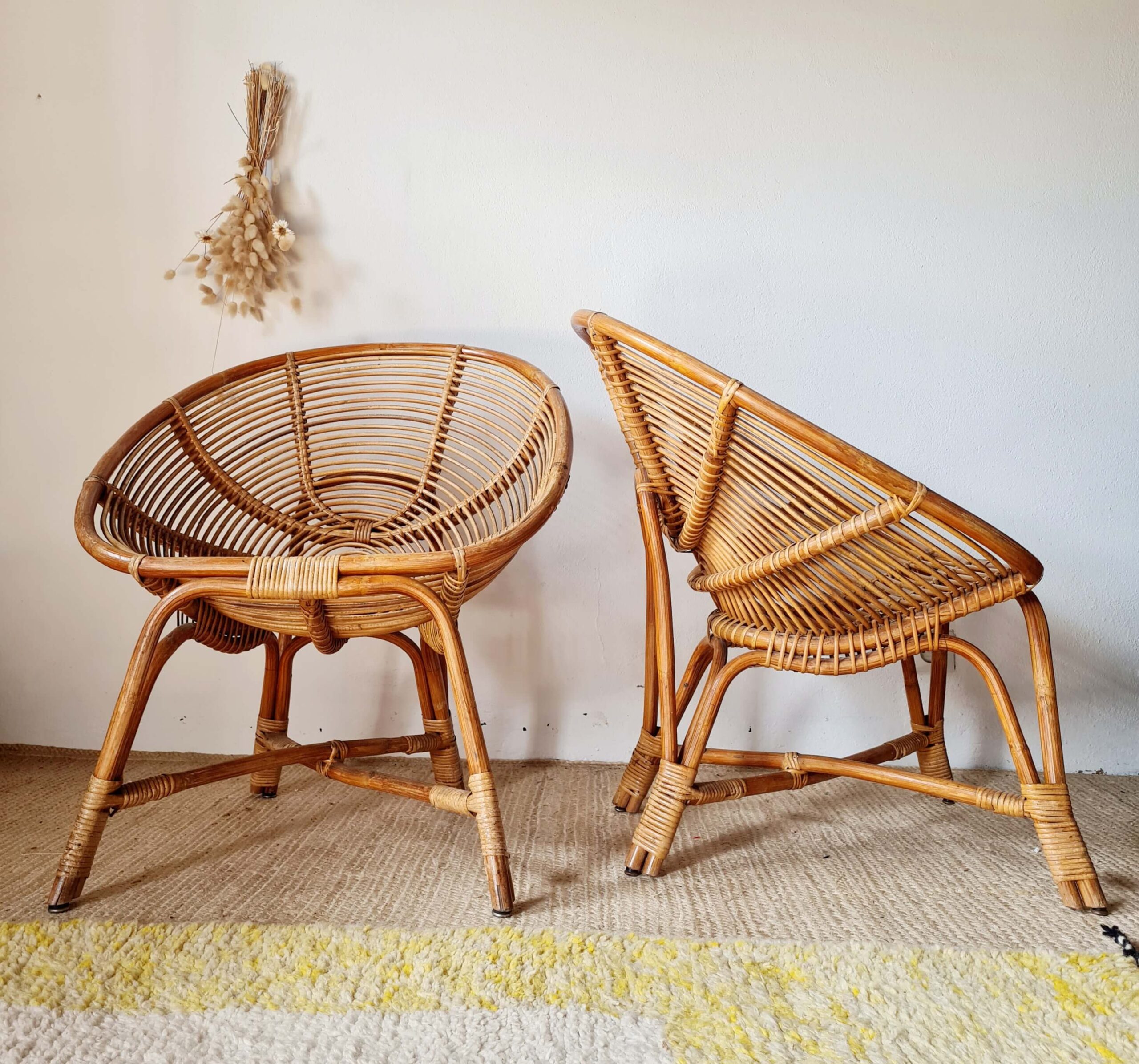 Deux fauteuils coquille en rotin tressé disposés côte à côte, décorés de fleurs séchées suspendues au mur.