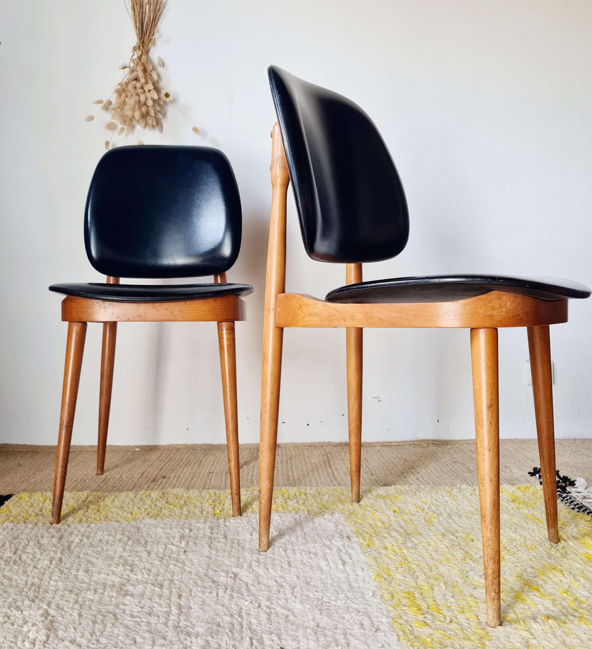 Deux chaises de style Mid-Century en bois clair et vinyle noir, l'une de face et l'autre de profil, installées sur un tapis de jute et de laine.