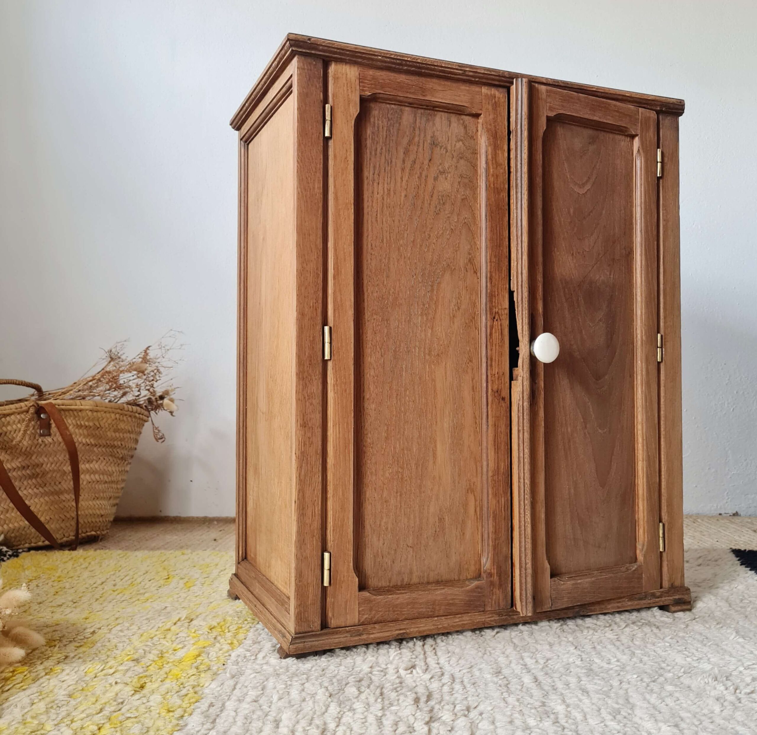 Petite armoire parisienne en bois brut fermée, posée sur un tapis, montrant ses deux portes moulurées et son bouton en céramique blanche.