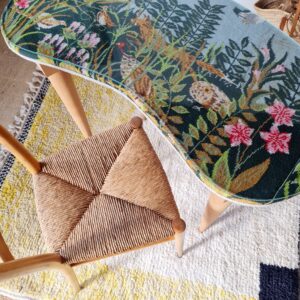 Vue aérienne d'une table basse haricot vintage et d'une chaise rétro avec assise en paille tressée, installées sur un tapis en laine épais blanc et jaune.