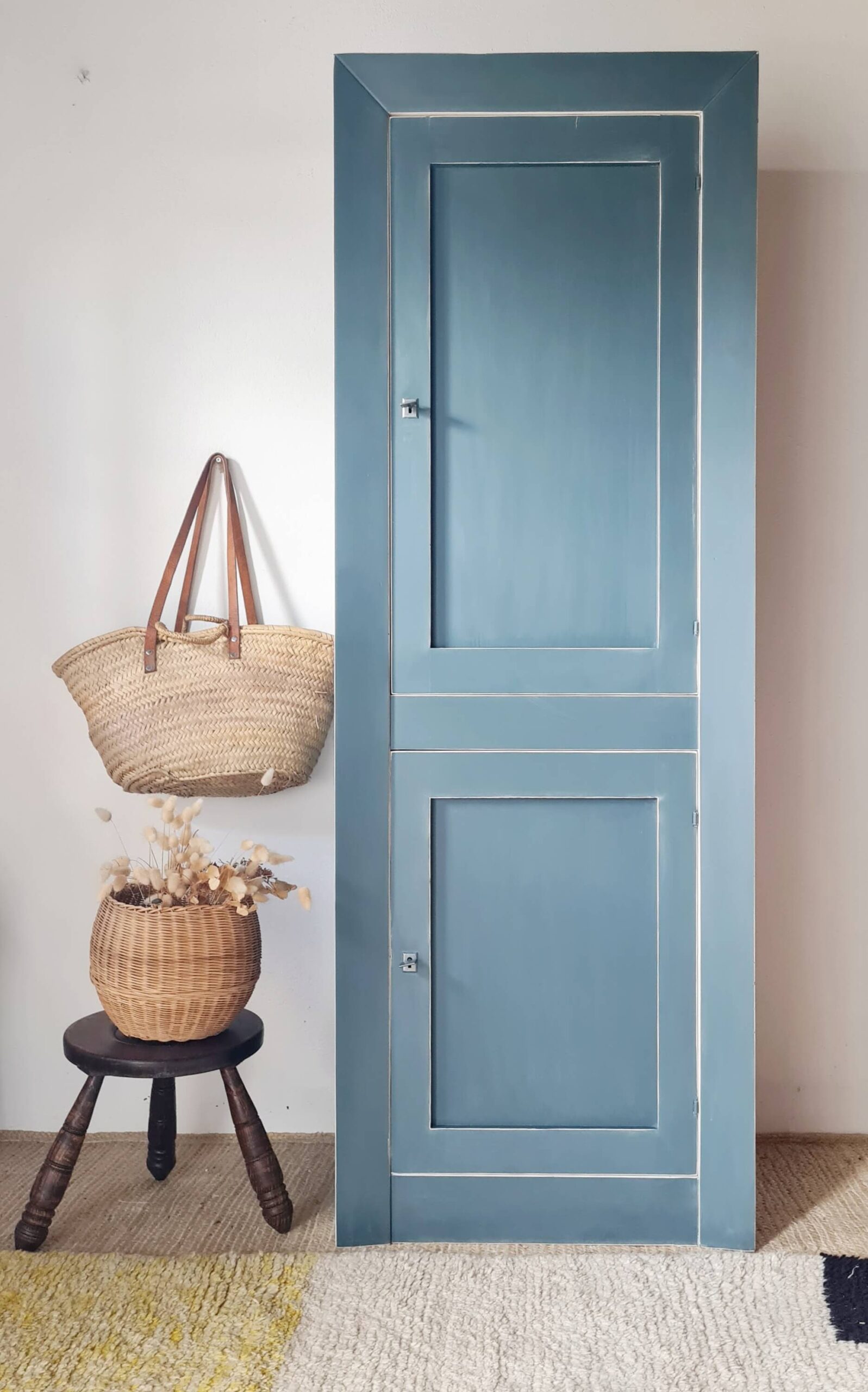 Armoire ancienne peinte en bleu avec finition vintage, accompagnée d'un panier en osier et d'un tabouret en bois dans un intérieur décoré.