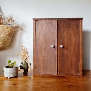 Photo de face d'une petite armoire à pharmacie vintage en bois marron avec ses deux portes fermées et ses boutons de tirage métalliques. Le meuble est mis en situation dans un intérieur lumineux au style bohème et rustique.