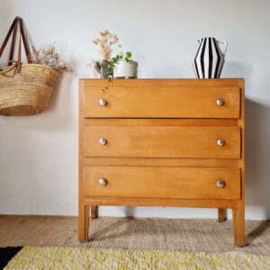 Commode vintage en bois clair à 3 tiroirs dans un intérieur rétro avec panier en osier et plantes