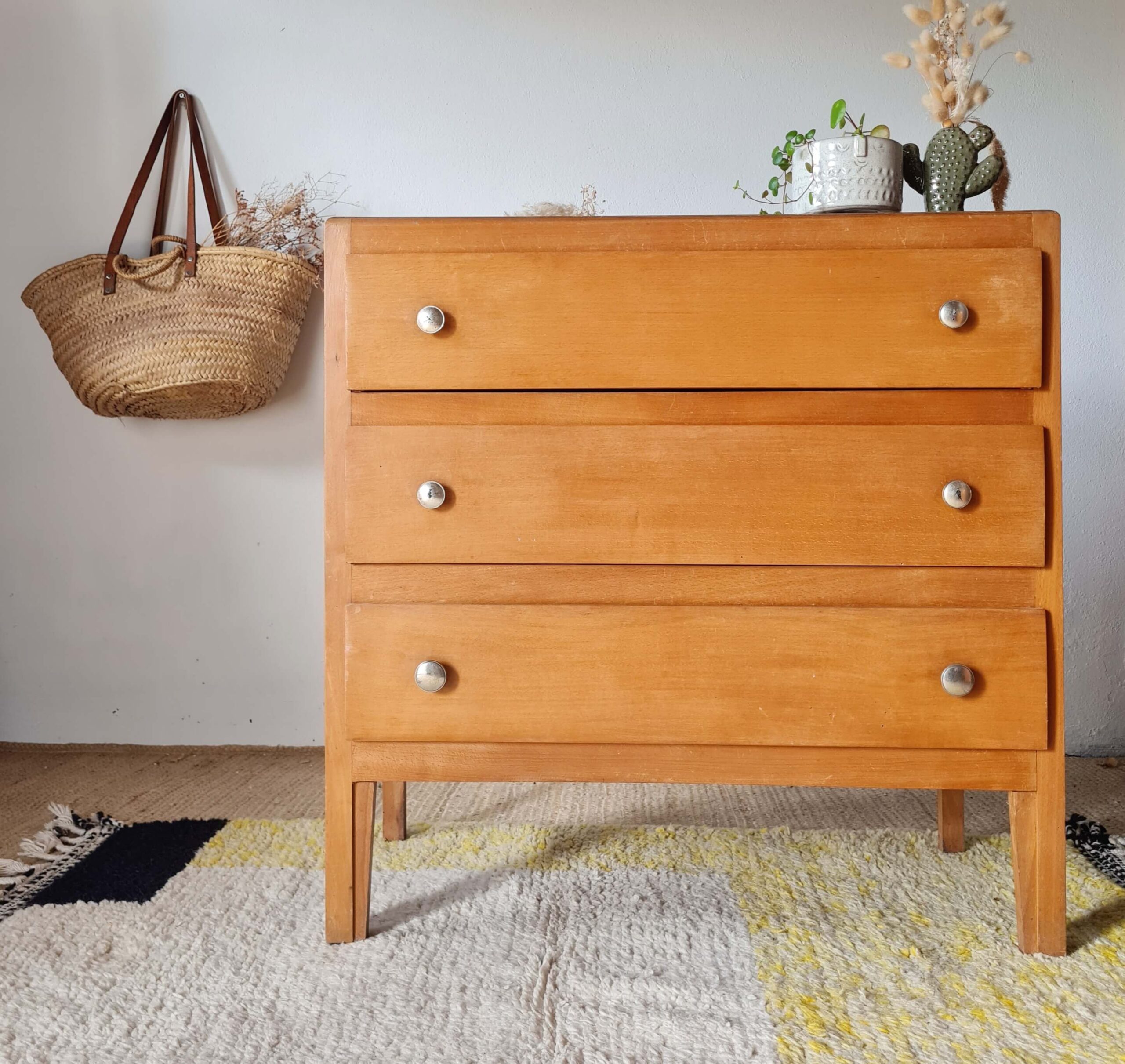 Vue de face d'une commode rétro en bois blond à trois tiroirs et pieds fuselés.