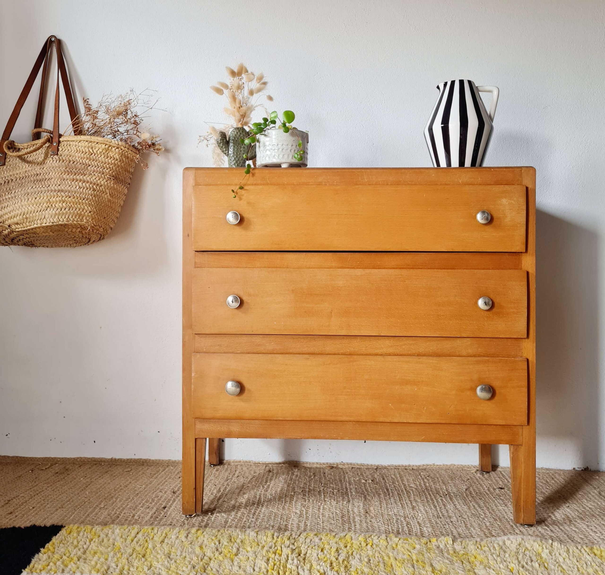 Commode vintage en bois clair à 3 tiroirs dans un intérieur rétro avec panier en osier et plantes