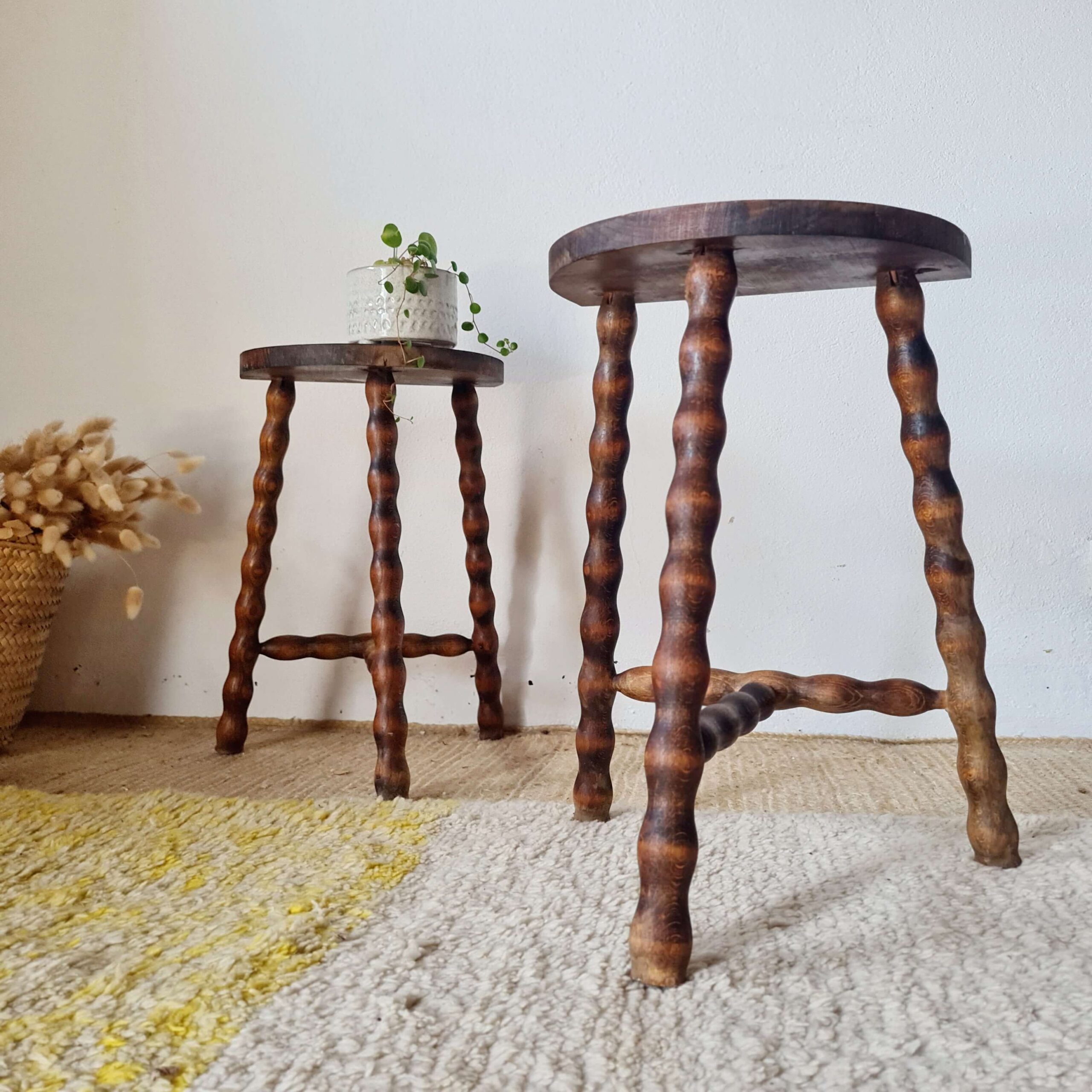 Vue en perspective de deux tabourets vintage en bois tourné posés sur un tapis en laine texturé, avec une petite plante en pot sur l'assise du fond.