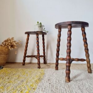 Mise en scène de deux tabourets anciens de style rustique en bois massif à pieds tournés, disposés de biais près d'un panier en osier rempli de fleurs séchées