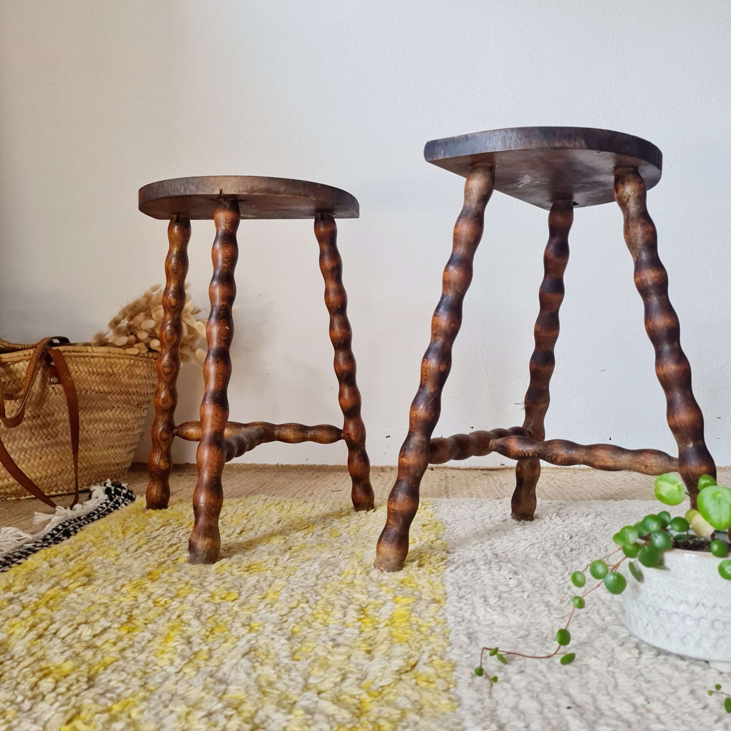 Décoration d'intérieur bohème présentant une paire de tabourets trépieds en bois massif avec un panier en osier en arrière-plan et une plante tombante au premier plan