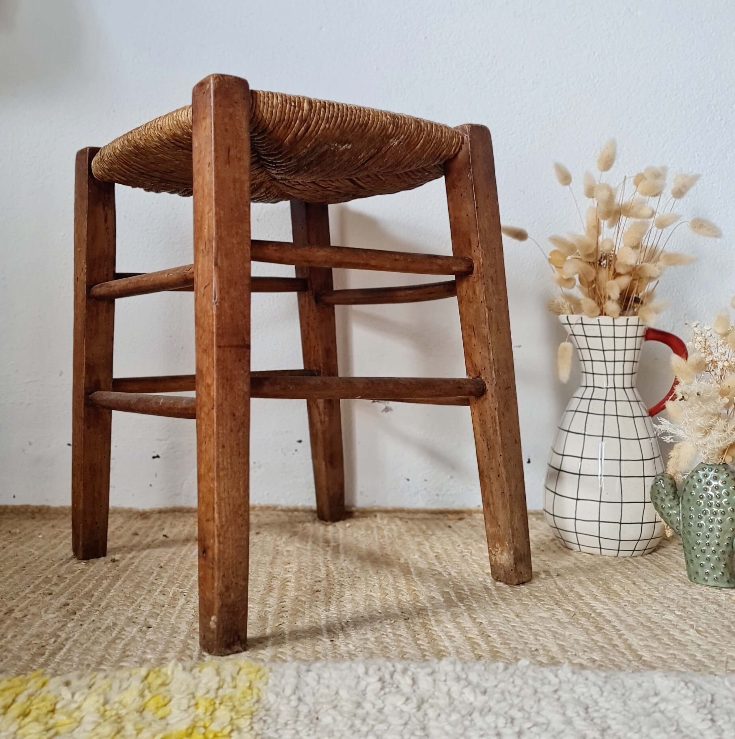 Petit tabouret rétro en bois massif et assise en paille à côté d'un vase blanc à carreaux noirs dans une ambiance chaleureuse.