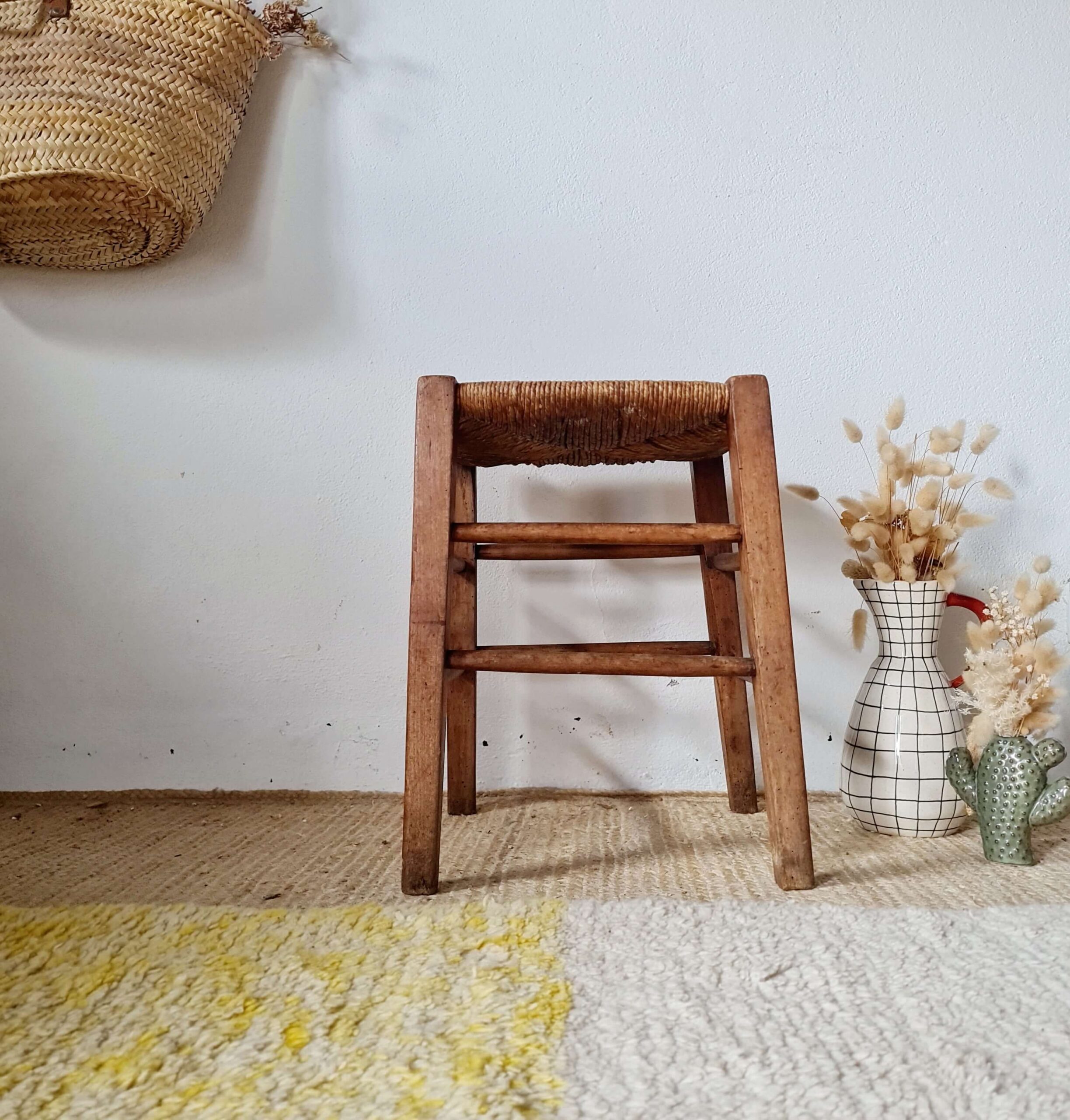 Vue d'ensemble d'un tabouret de ferme ancien en bois brut et paillage traditionnel, posé sur un tapis en sisal dans une décoration bohème chic.