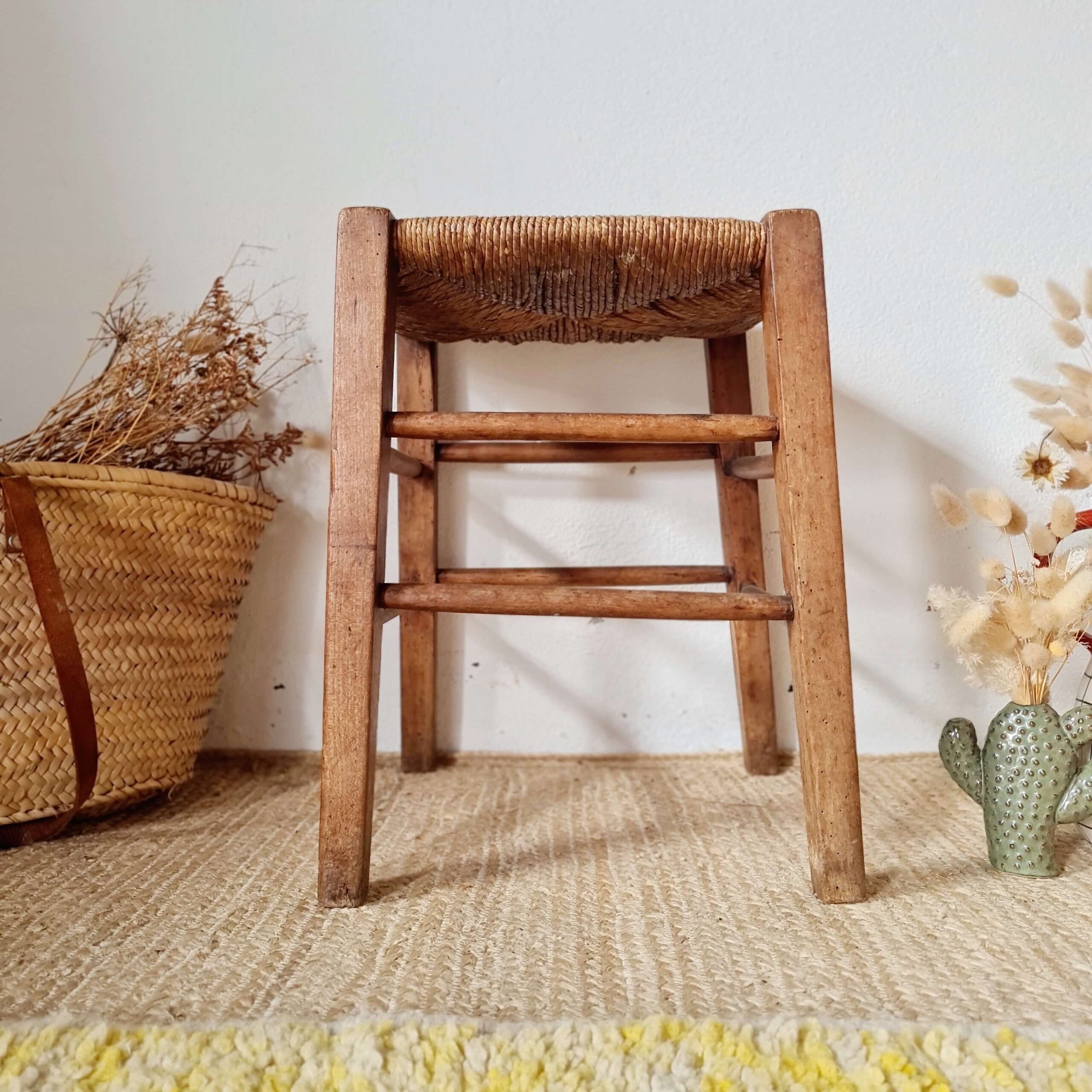 Tabouret ancien en bois et paille tressée, photographié de face dans une ambiance bohème avec un panier en osier à gauche et un vase cactus à droite.