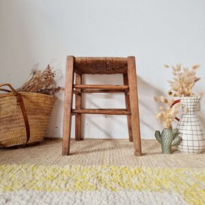 Vue de face d'un tabouret rustique en bois et paille, posé sur un tapis texturé, entouré d'un panier en osier et d'un vase quadrillé avec des fleurs séchées.