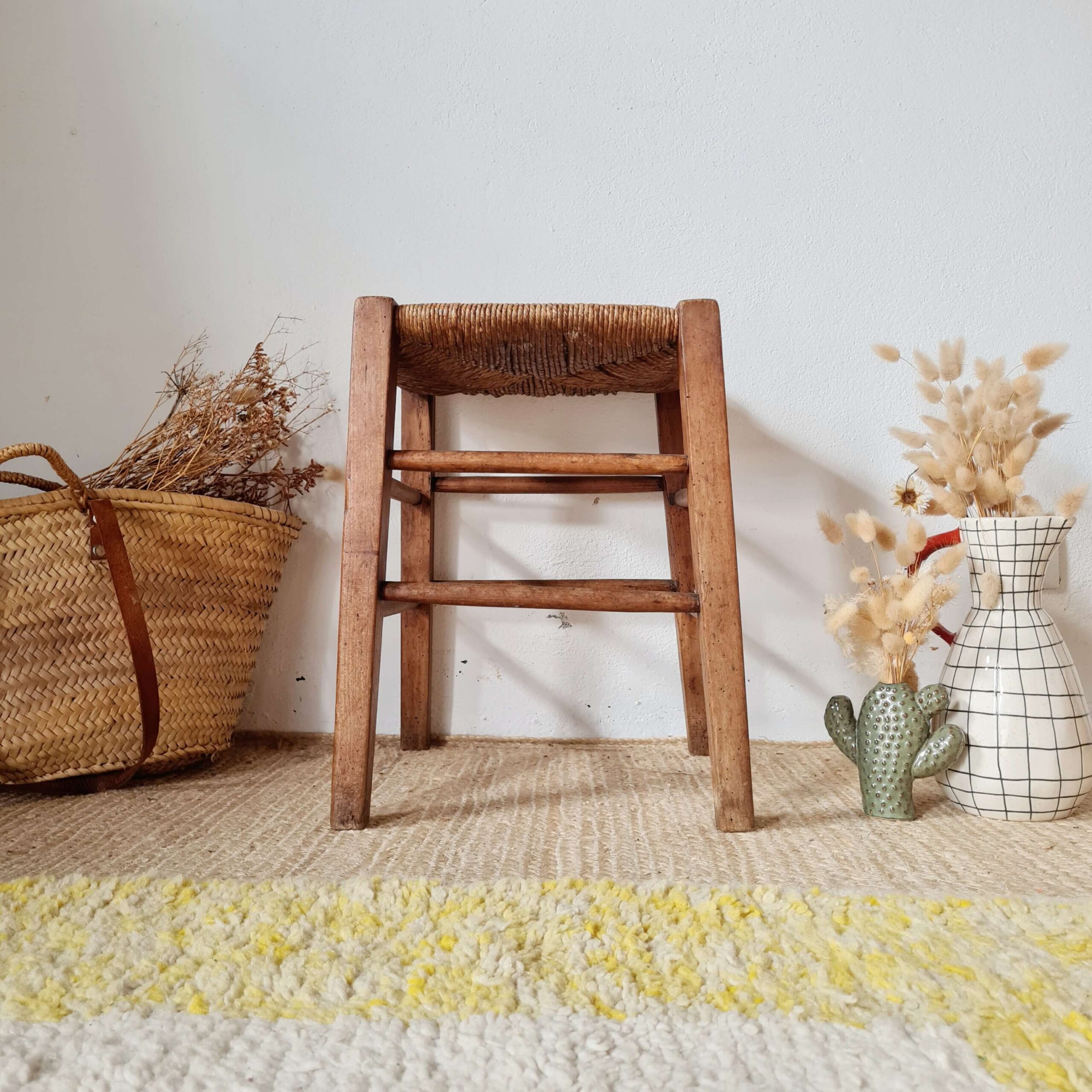 Vue de face d'un tabouret rustique en bois et paille, posé sur un tapis texturé, entouré d'un panier en osier et d'un vase quadrillé avec des fleurs séchées.