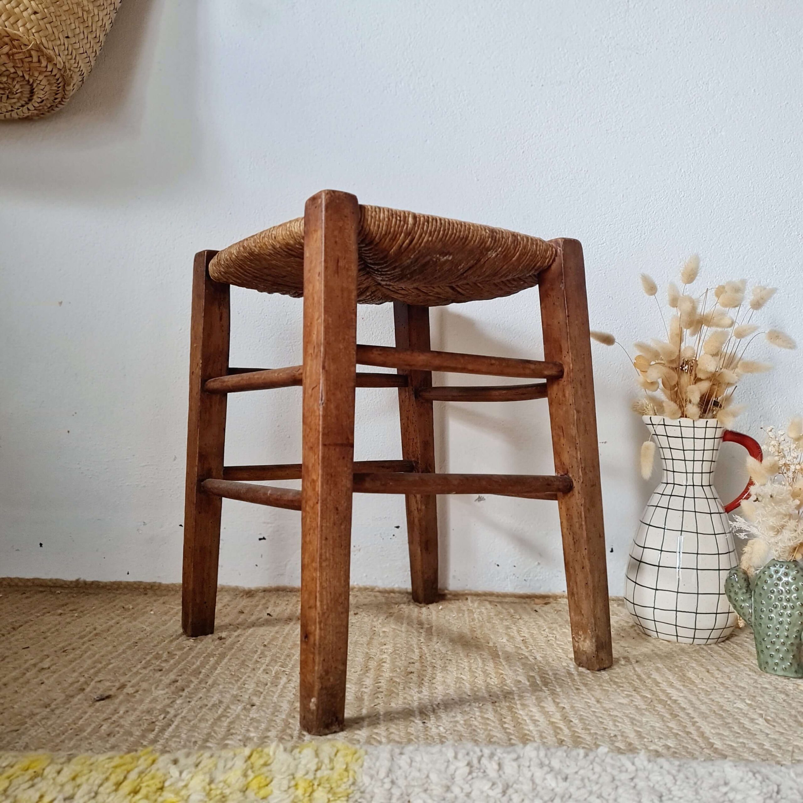 Un tabouret rustique vintage en bois et paille avec une belle patine, photographié de face à côté d'un vase quadrillé rempli de fleurs séchées.
