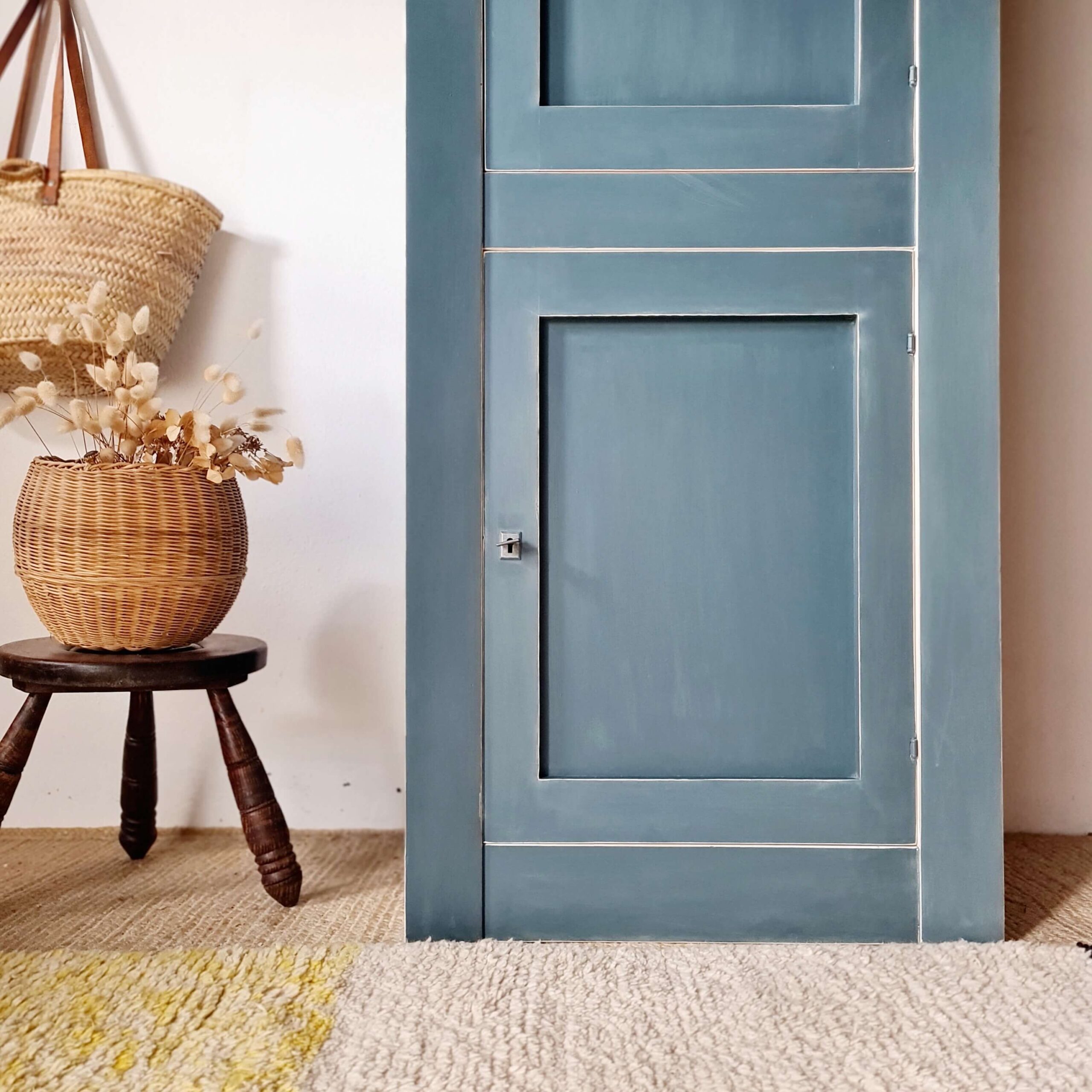 Gros plan sur la porte inférieure d'une armoire bleue relookée, avec un panier rempli de fleurs séchées en arrière-plan