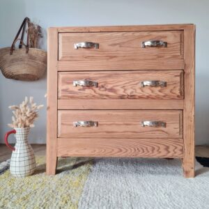 Commode vintage à trois tiroirs en bois de pin massif avec poignées en métal, décorée d'un vase et d'un panier en osier suspendu au mur.