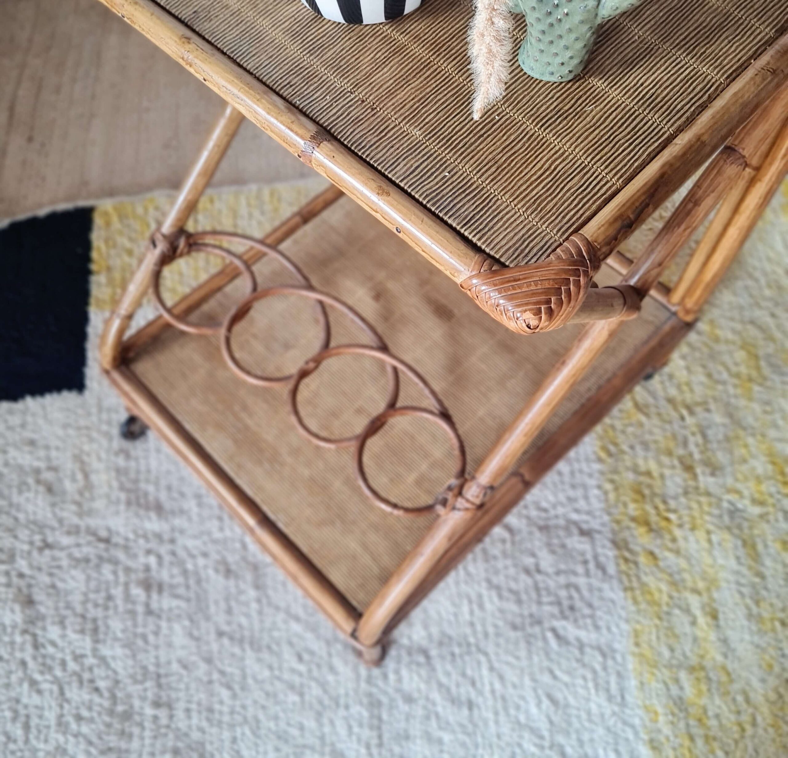 Vue en plongée d'une desserte de bar vintage en rotin avec porte-bouteilles intégré et étagère en cannage.