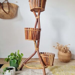 Vue d'ensemble en plan moyen d'un grand porte-plante vintage en bambou à trois étages, avec ses pots garnis de plantes, un panier en osier de fleurs séchées à droite et deux petits pots au sol à gauche.
