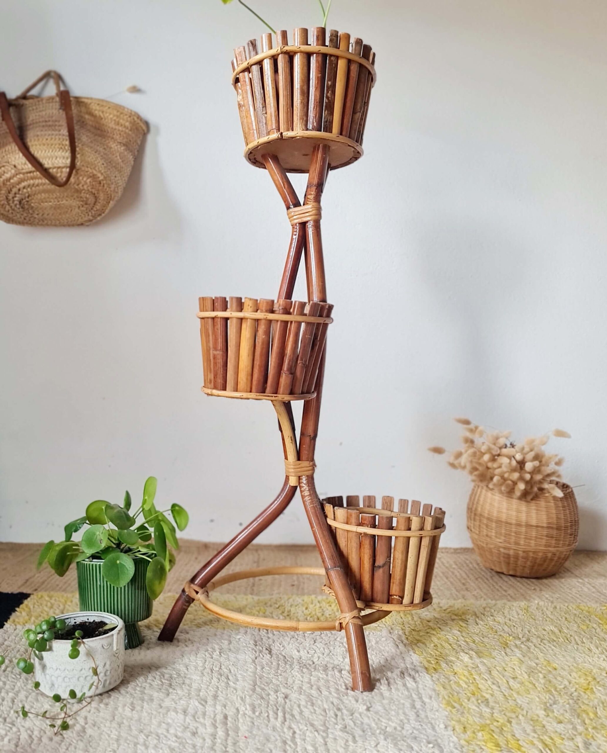 Vue d'ensemble en plan moyen d'un grand porte-plante vintage en bambou à trois étages, avec ses pots garnis de plantes, un panier en osier de fleurs séchées à droite et deux petits pots au sol à gauche.