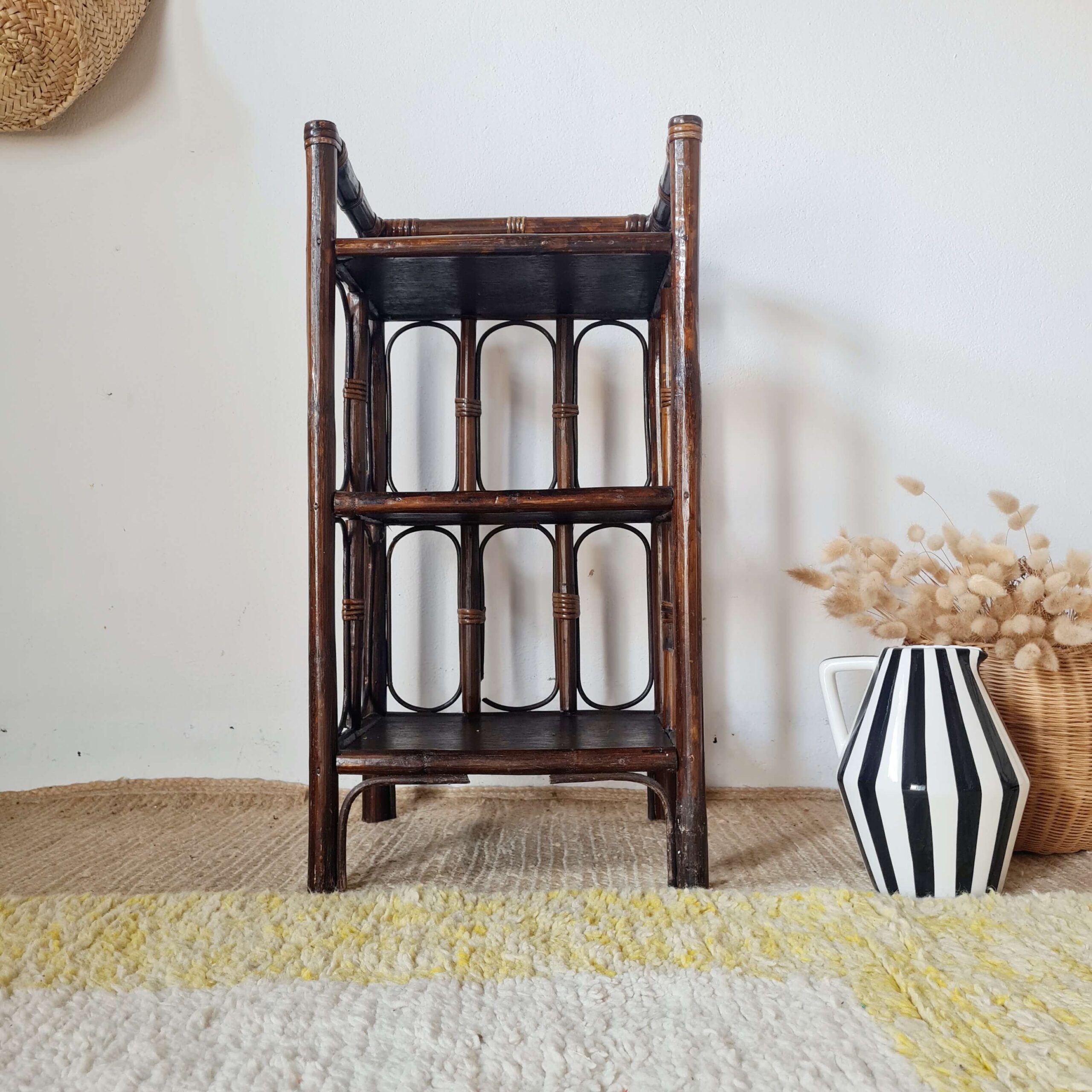 Étagère vintage en rotin brun foncé à trois niveaux, présentée avec un pichet rayé noir et blanc.