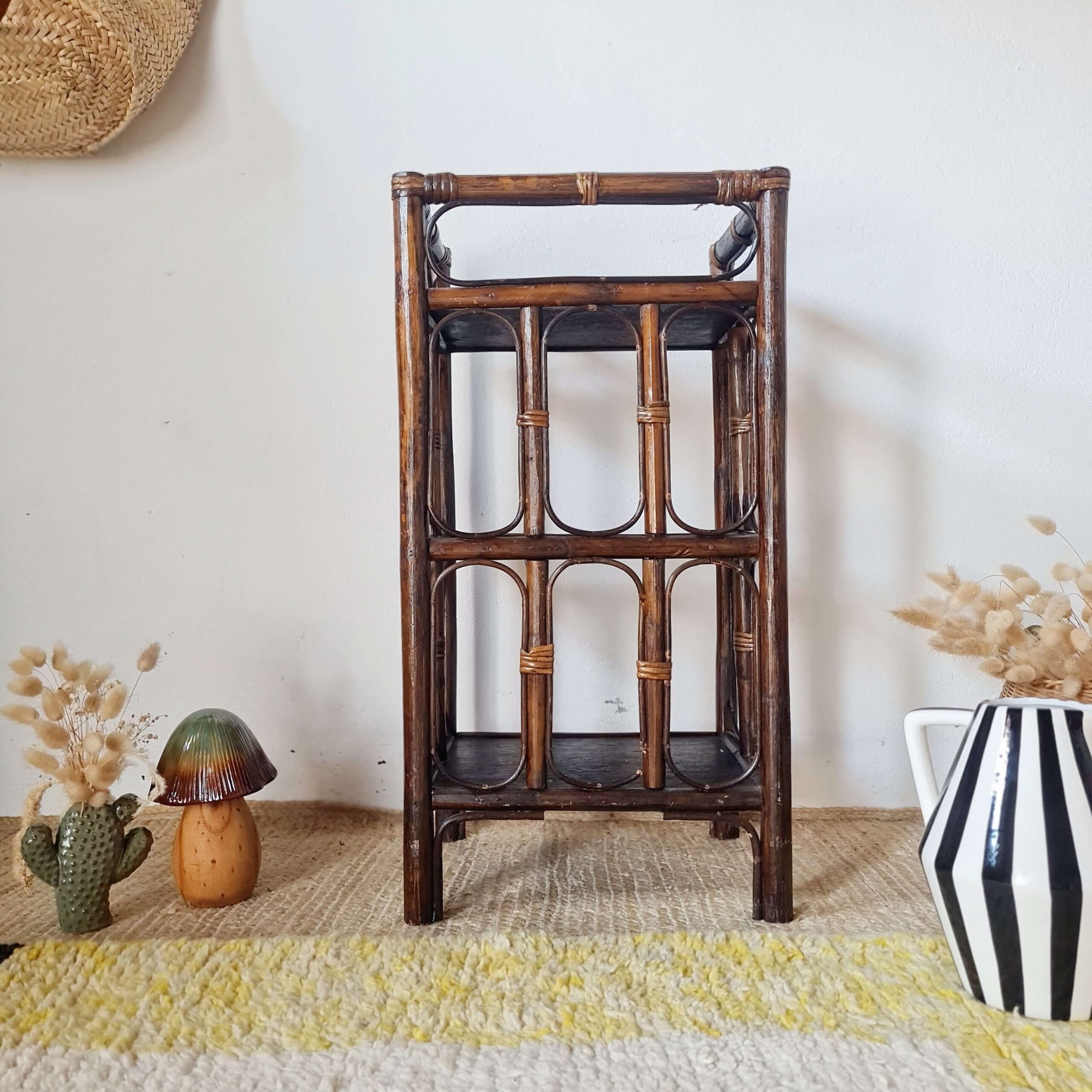 Vue de face d'une petite étagère de rangement en rotin foncé à plusieurs plateaux sur un tapis clair.