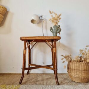 Petite table basse ou guéridon ancien en rotin et bambou, décoré d'une lampe vintage et d'un vase en forme de cactus, sur fond de mur blanc.