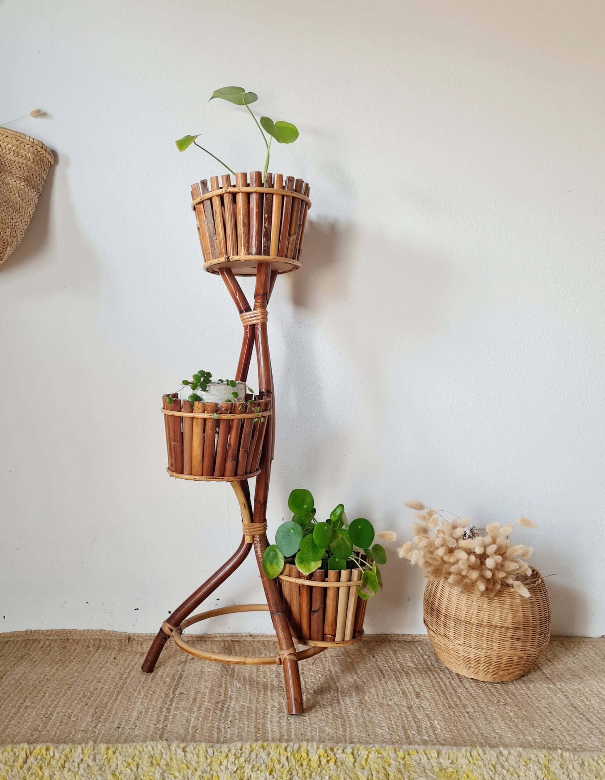 Vue d'ensemble d'un grand porte-plante vintage en bambou et rotin à trois niveaux, accueillant différentes plantes vertes, avec un panier en osier rempli de fleurs séchées posé au sol contre un mur blanc.