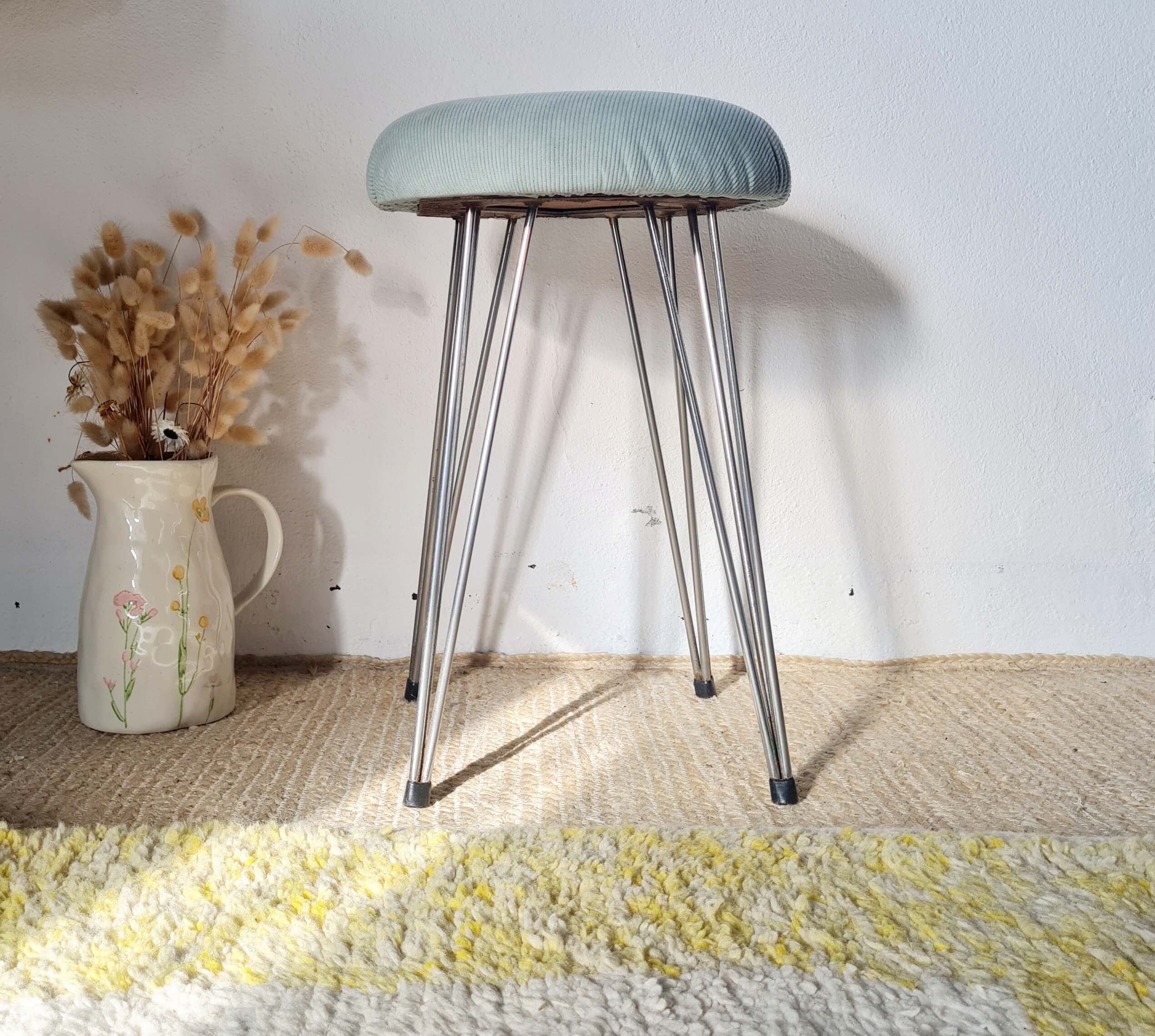 Tabouret vintage à assise ronde en velours côtelé bleu clair et pieds compas en métal, posé sur un tapis texturé.