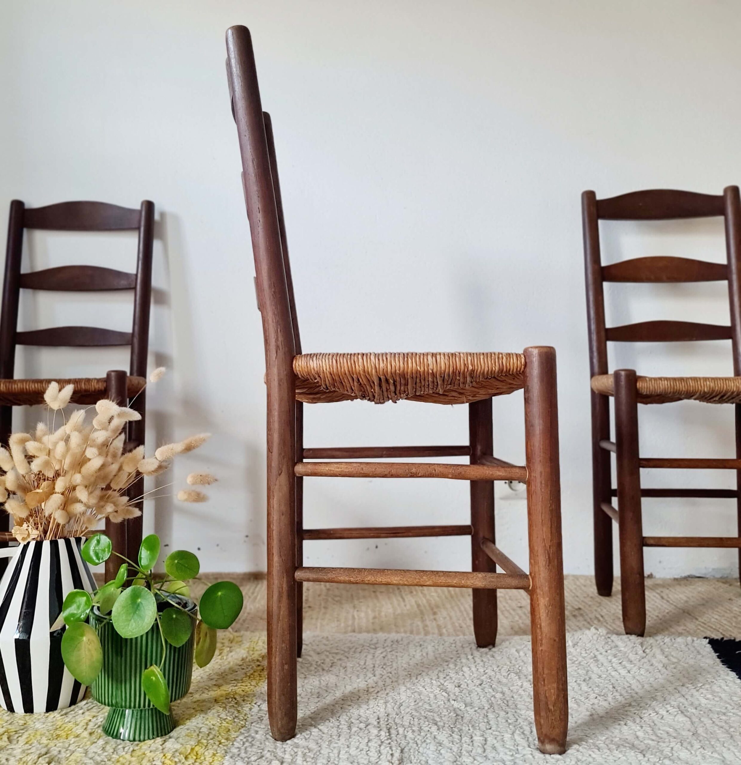Profil d'une chaise de style rustique en bois tourné et dossier à barreaux, assise en paille tressée visible, sur un tapis clair contre un mur blanc