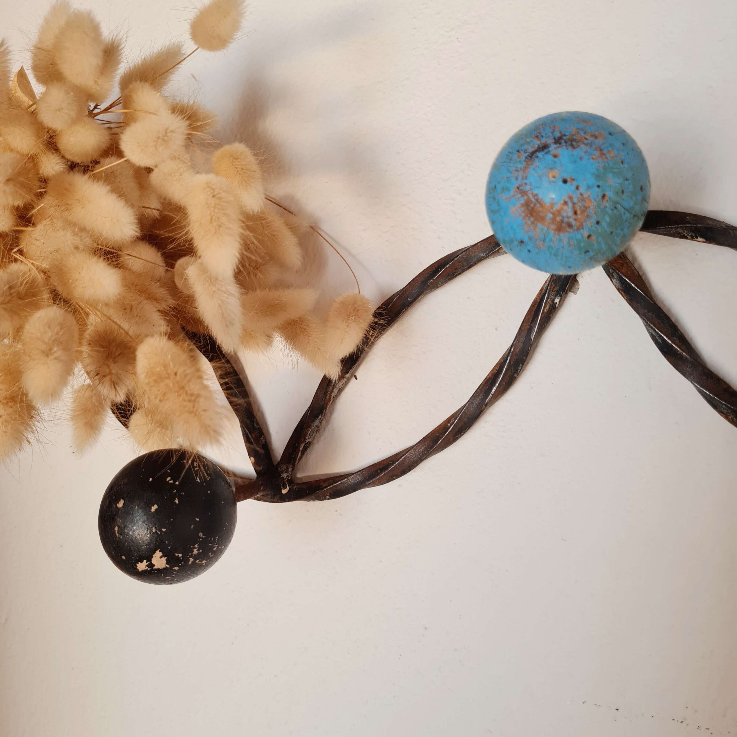 Gros plan sur la boule noire inférieure et la boule bleue supérieure reliées par les torsades de métal foncé d'un porte-manteau rétro, avec des fleurs séchées floutées en arrière-plan.