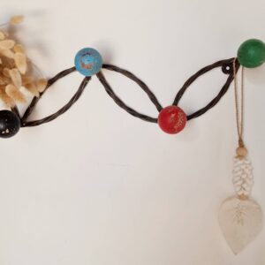 Vue de face d'un porte-manteau mural en métal torsadé foncé orné de boules en bois colorées (noir, bleu, rouge, vert), fixé sur un mur blanc avec un petit bouquet de fleurs séchées à gauche et une suspension macramé en forme de feuille à droite.