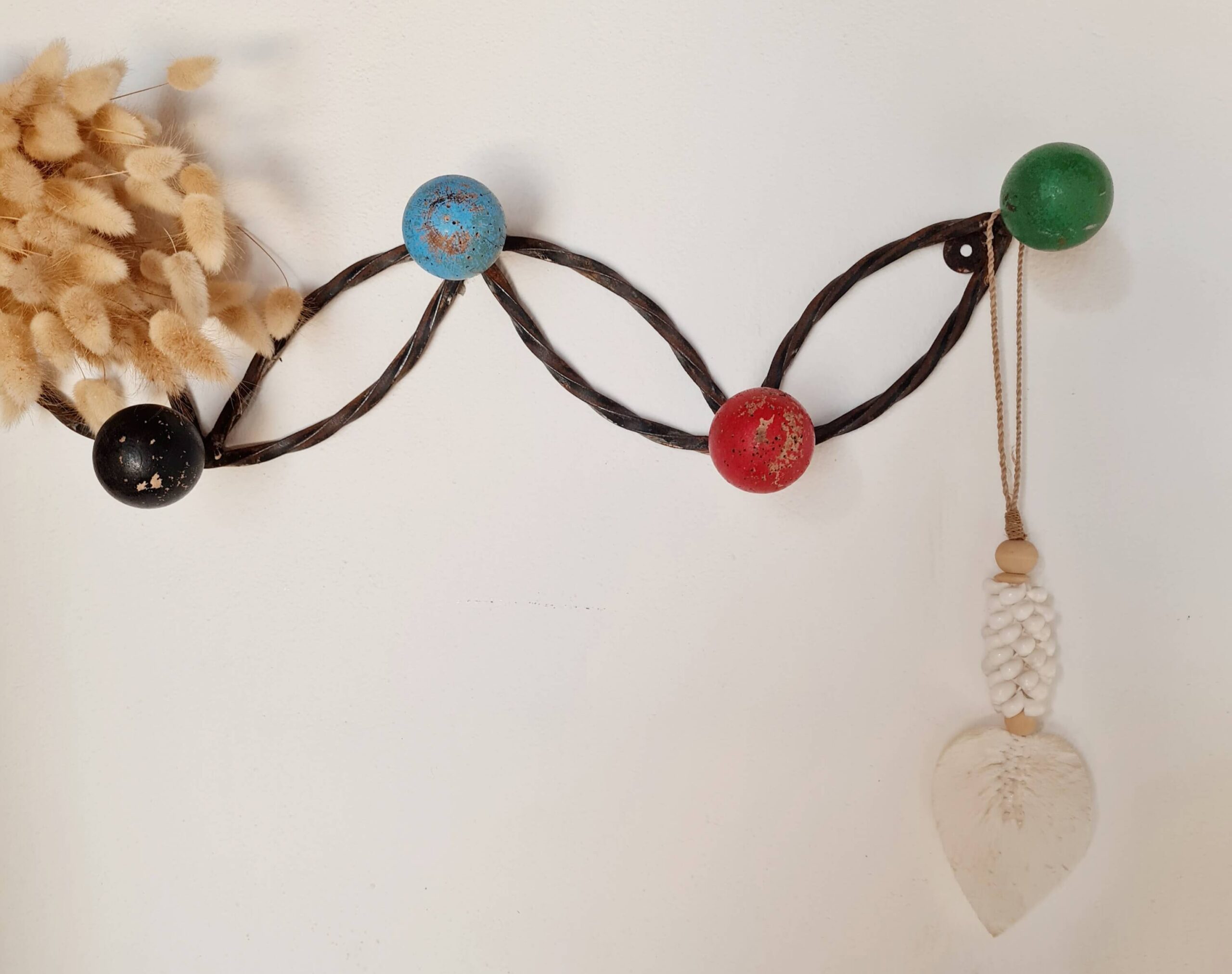 Vue de face d'un porte-manteau mural en métal torsadé foncé orné de boules en bois colorées (noir, bleu, rouge, vert), fixé sur un mur blanc avec un petit bouquet de fleurs séchées à gauche et une suspension macramé en forme de feuille à droite.