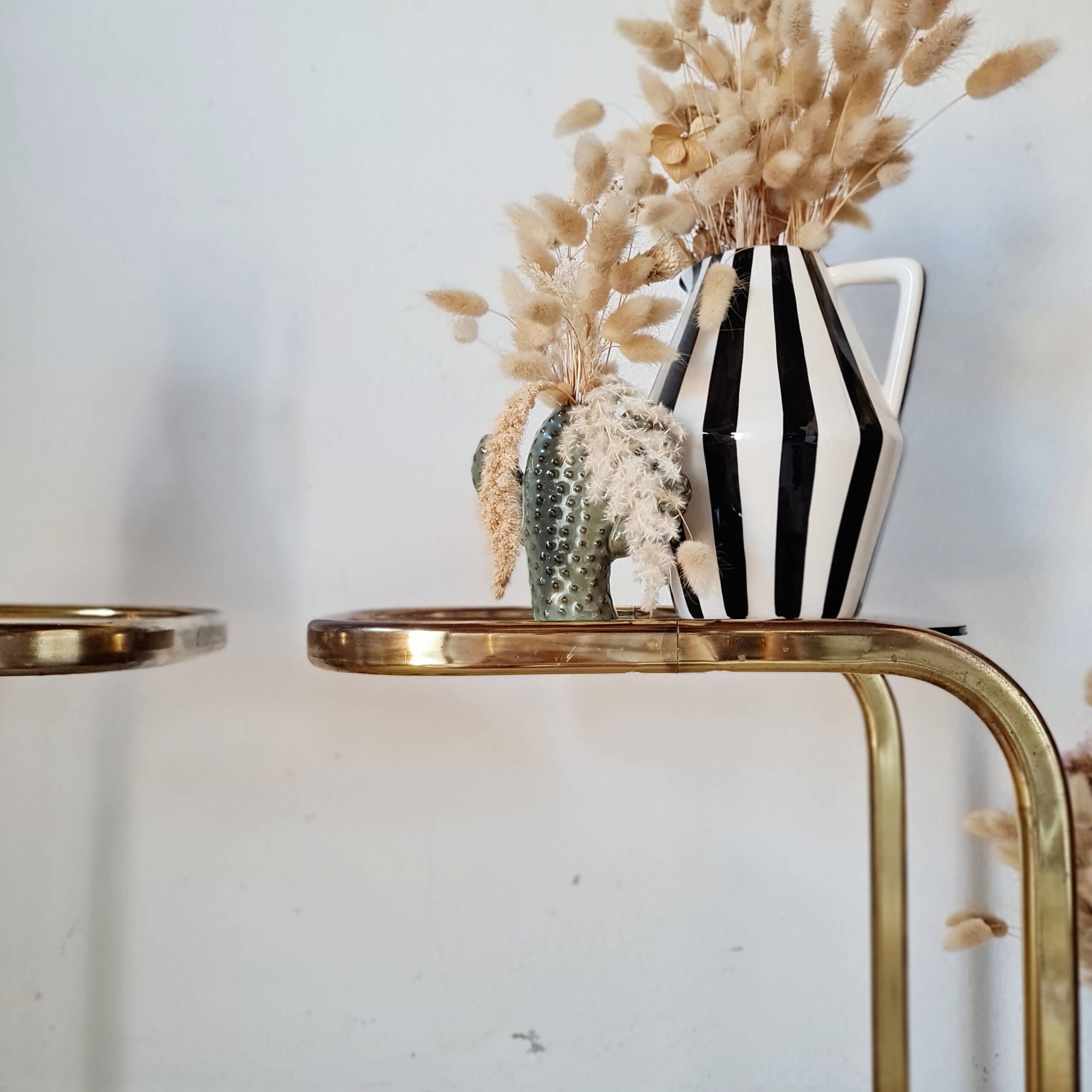 Gros plan sur l'une des tables de bout de canapé en C, structure en métal doré patiné et plateau en verre fuméun vase rayé noir et blanc.