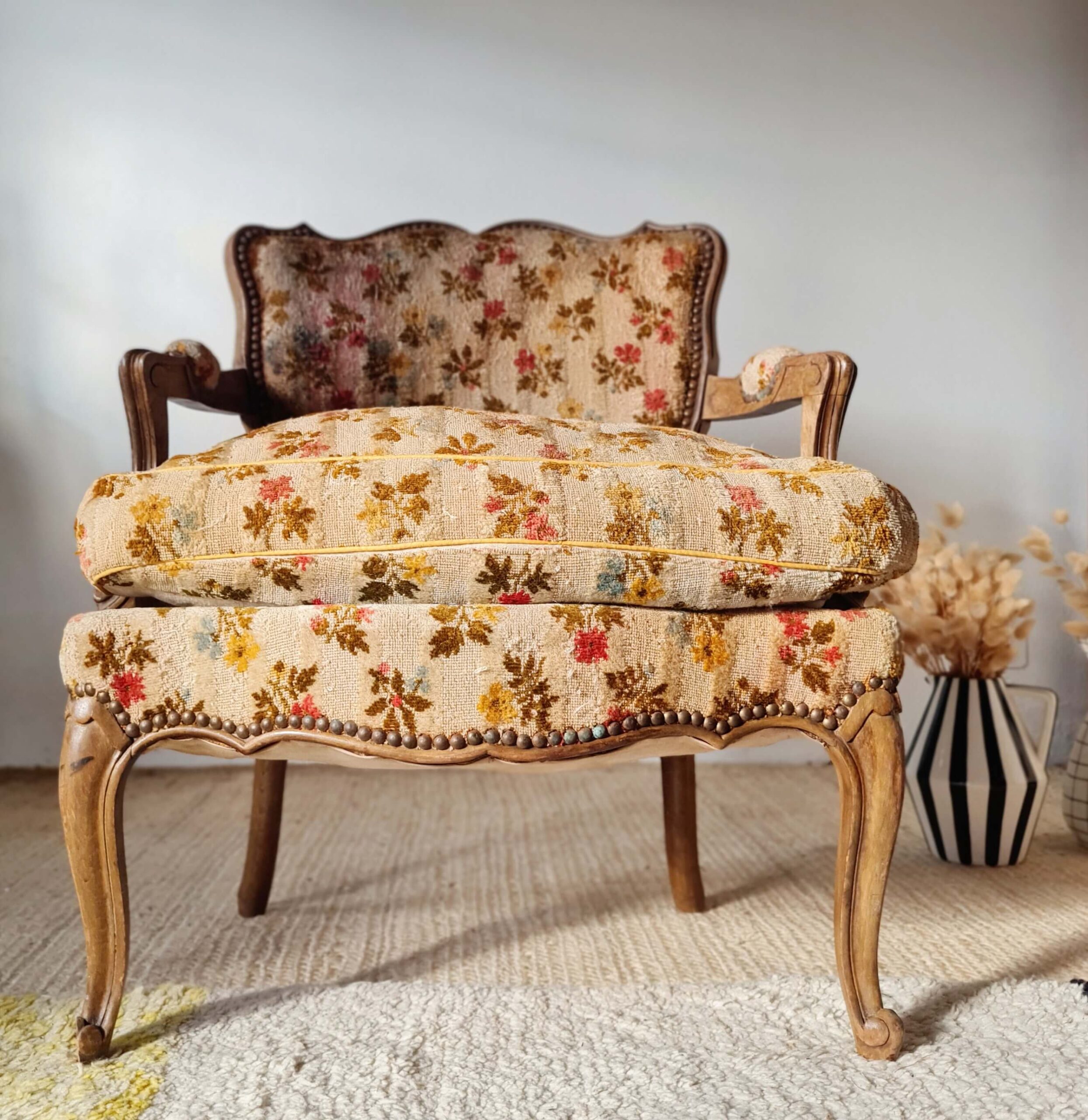 Vue de face d'un fauteuil crapaud vintage avec structure en bois sculpté et garniture cloutée, recouvert d'un tissu tapissier à motifs floraux. Il est posé sur un tapis en jonc de mer à côté d'un vase rayé noir et blanc rempli de fleurs séchées.