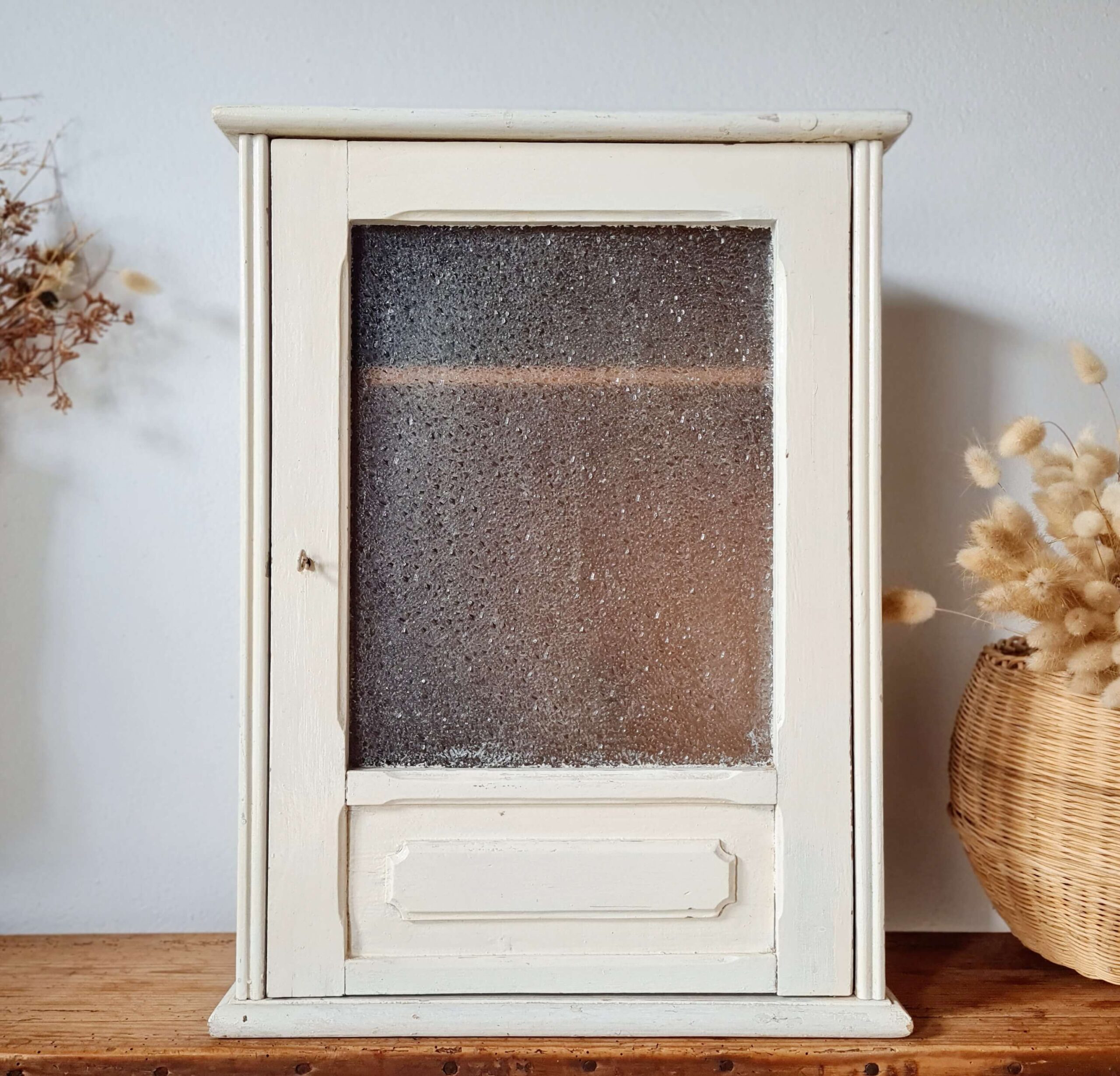 Autre prise de vue de face de la petite vitrine à pharmacie murale ancienne, mettant en valeur sa porte battante dotée d'un verre texturé et son corps en bois clair.