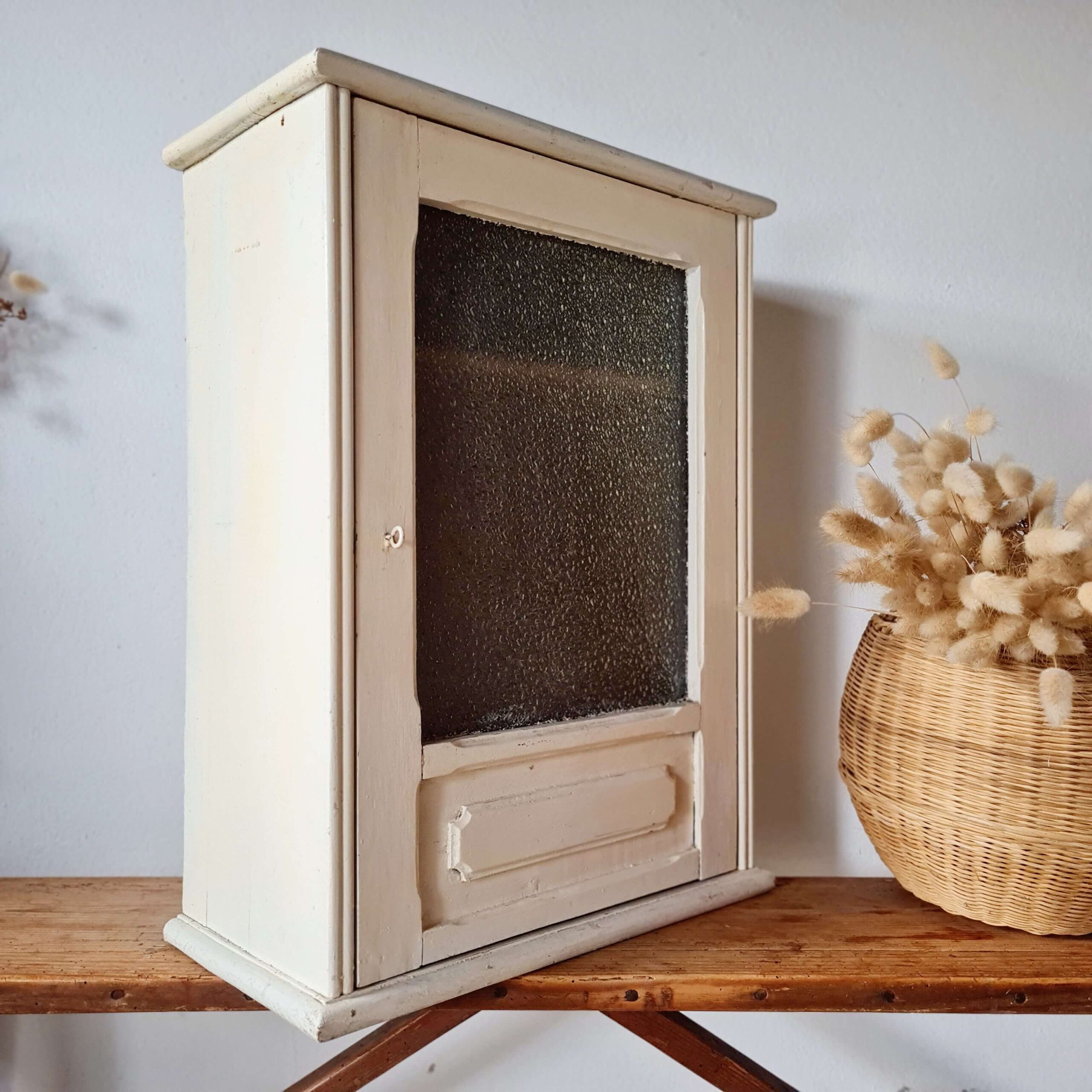 Vue de profil droit de la petite vitrine murale en bois blanc cassé, posée sur un établi en bois avec un panier d'herbes séchées à l'arrière-plan.