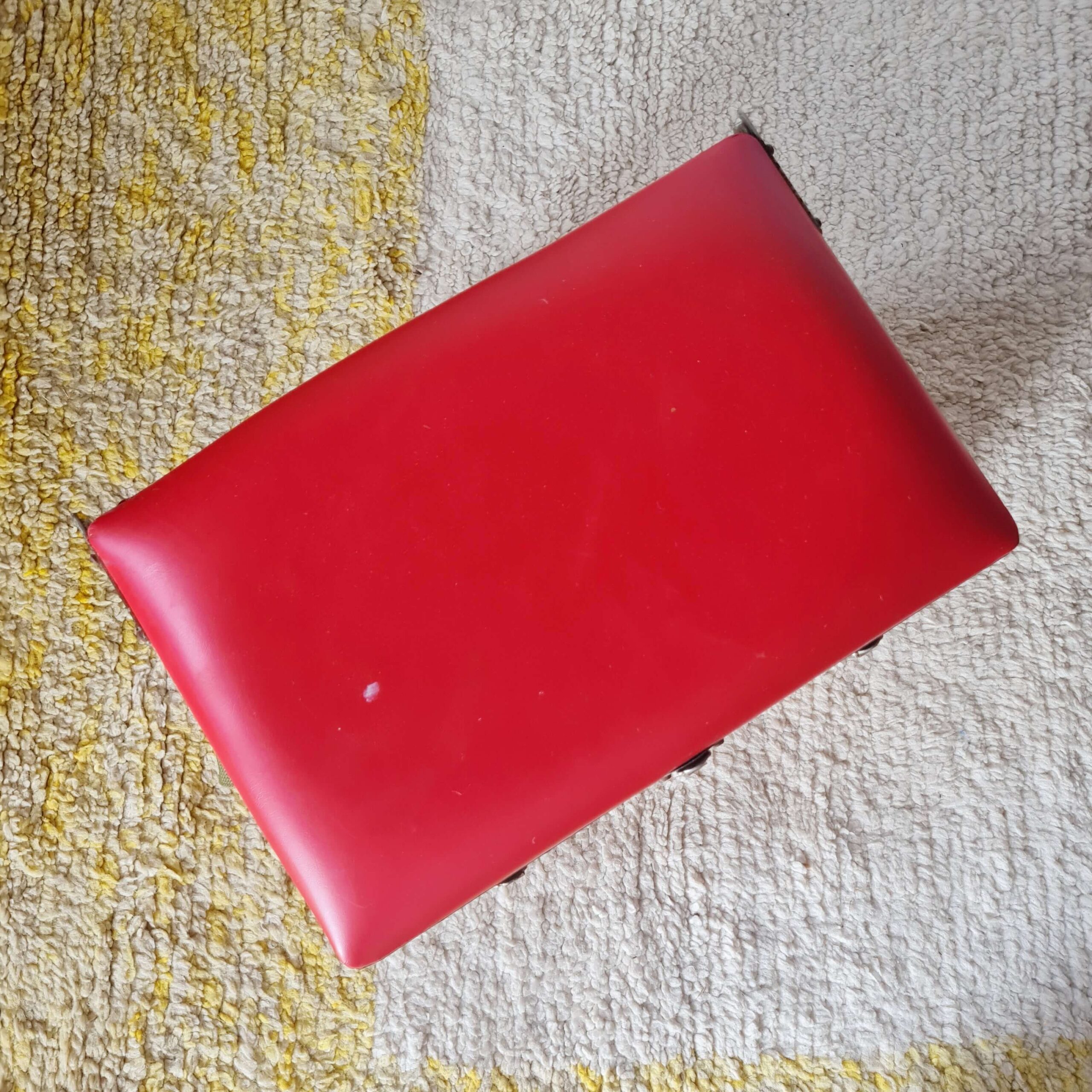 Plan zénithal montrant le dessus de l'assise rectangulaire en simili cuir rouge de l'ancien tabouret de pêcheur. Le revêtement est lisse avec quelques traces d'usage.