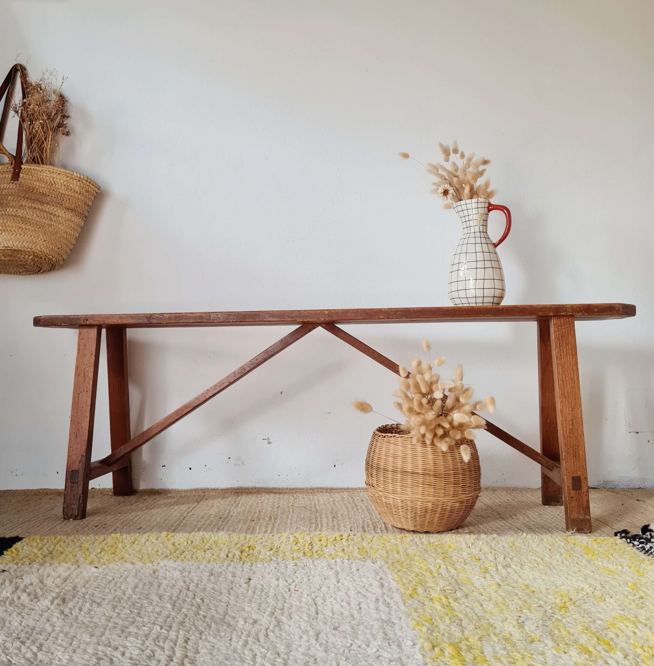 Banc ancien en bois massif patiné de style ferme, posé sur un tapis berbère avec en arrière-plan un mur blanc, un panier en osier