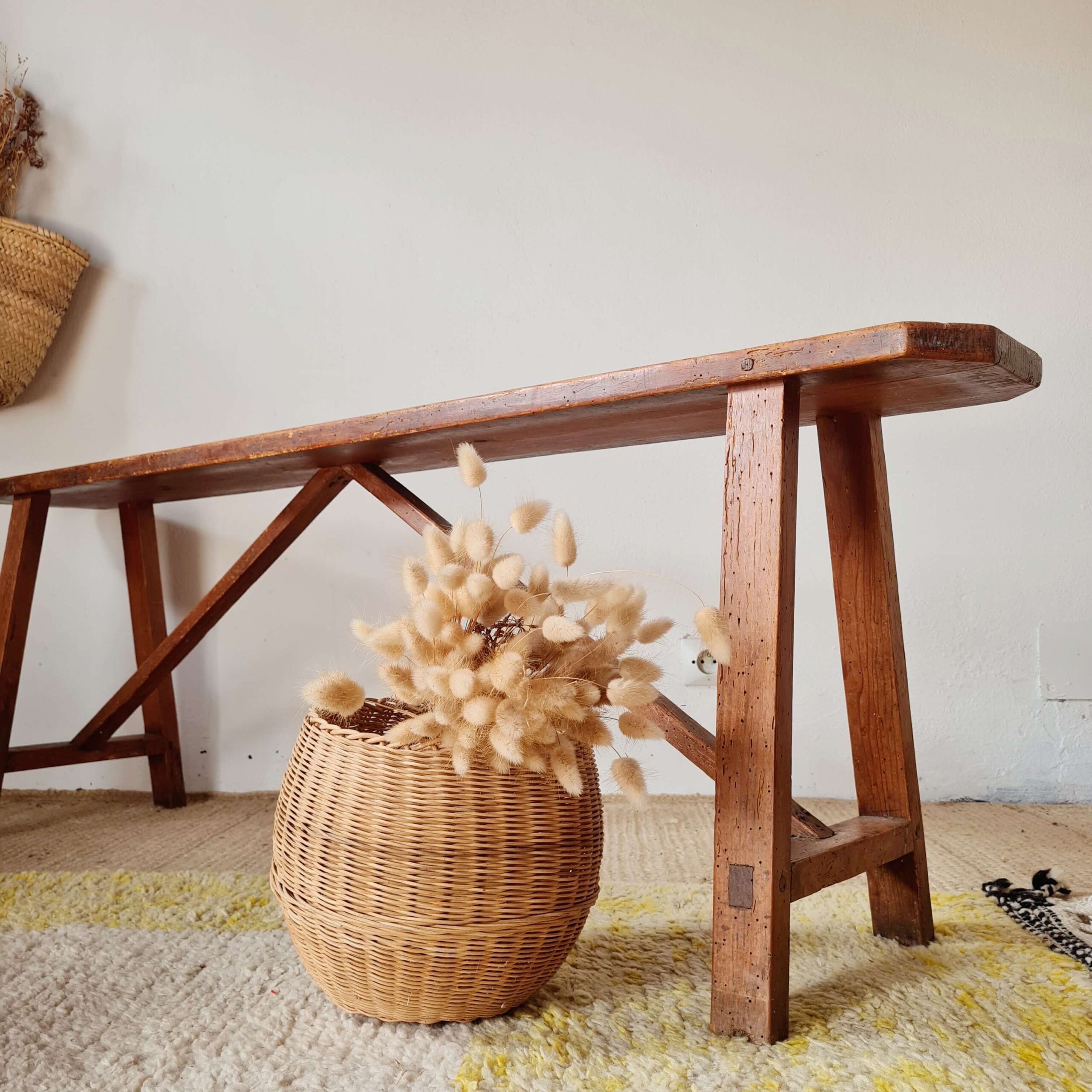 Banc ancien en bois massif patiné de style ferme, posé sur un tapis berbère avec en arrière-plan un mur blanc, un panier en osier