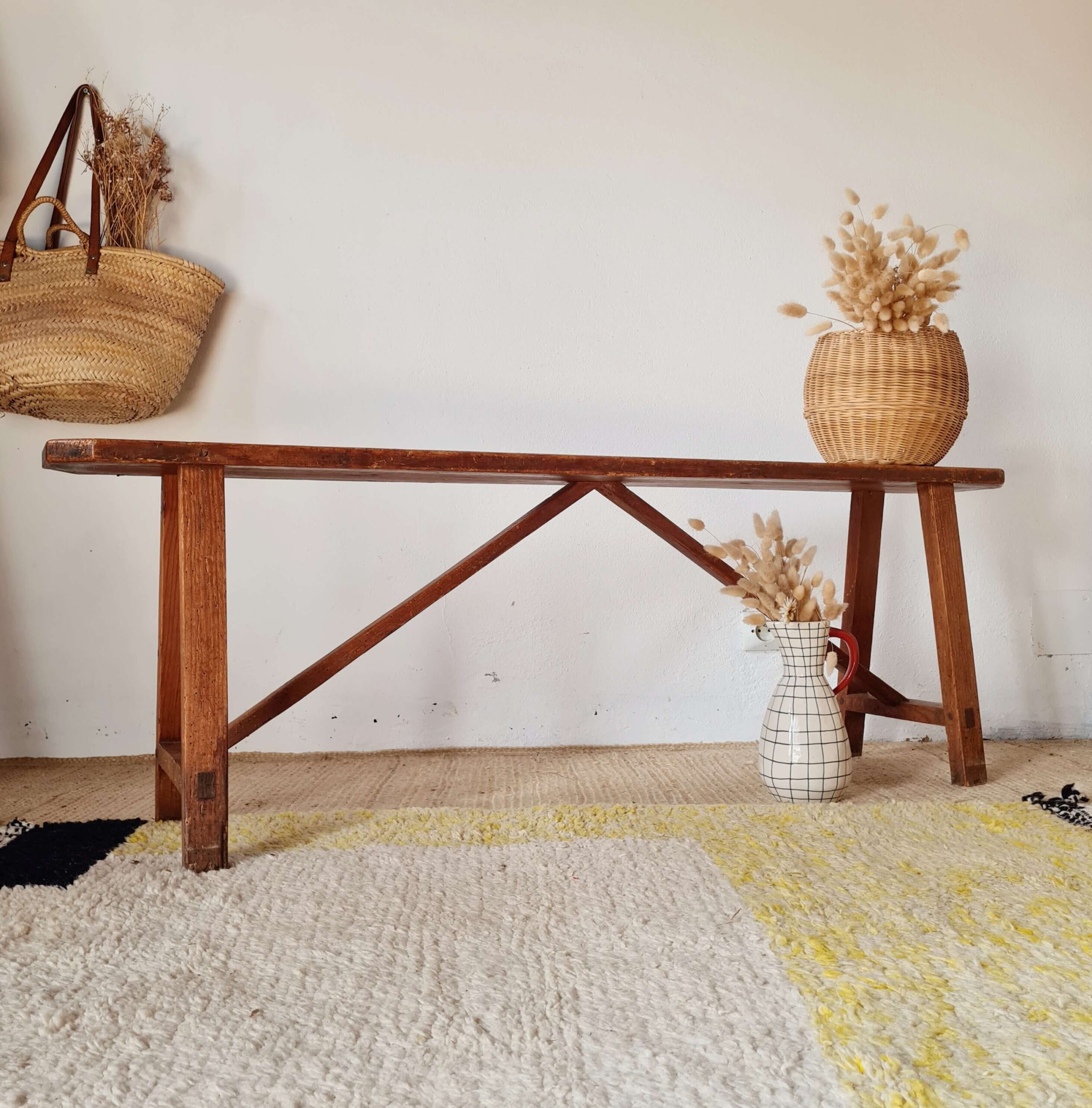 Banc ancien en bois massif patiné de style ferme, posé sur un tapis berbère avec en arrière-plan un mur blanc, un panier en osier