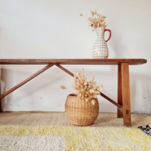 Banc ancien en bois massif patiné de style ferme, posé sur un tapis berbère avec en arrière-plan un mur blanc, un panier en osier