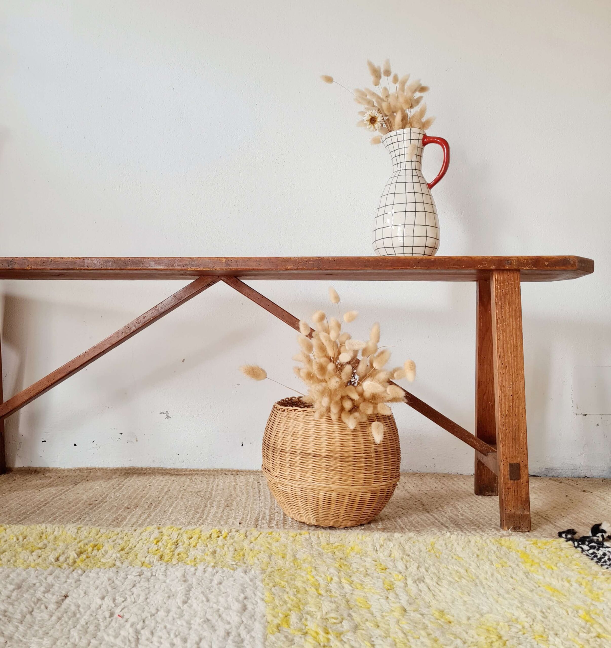 Banc ancien en bois massif patiné de style ferme, posé sur un tapis berbère avec en arrière-plan un mur blanc, un panier en osier