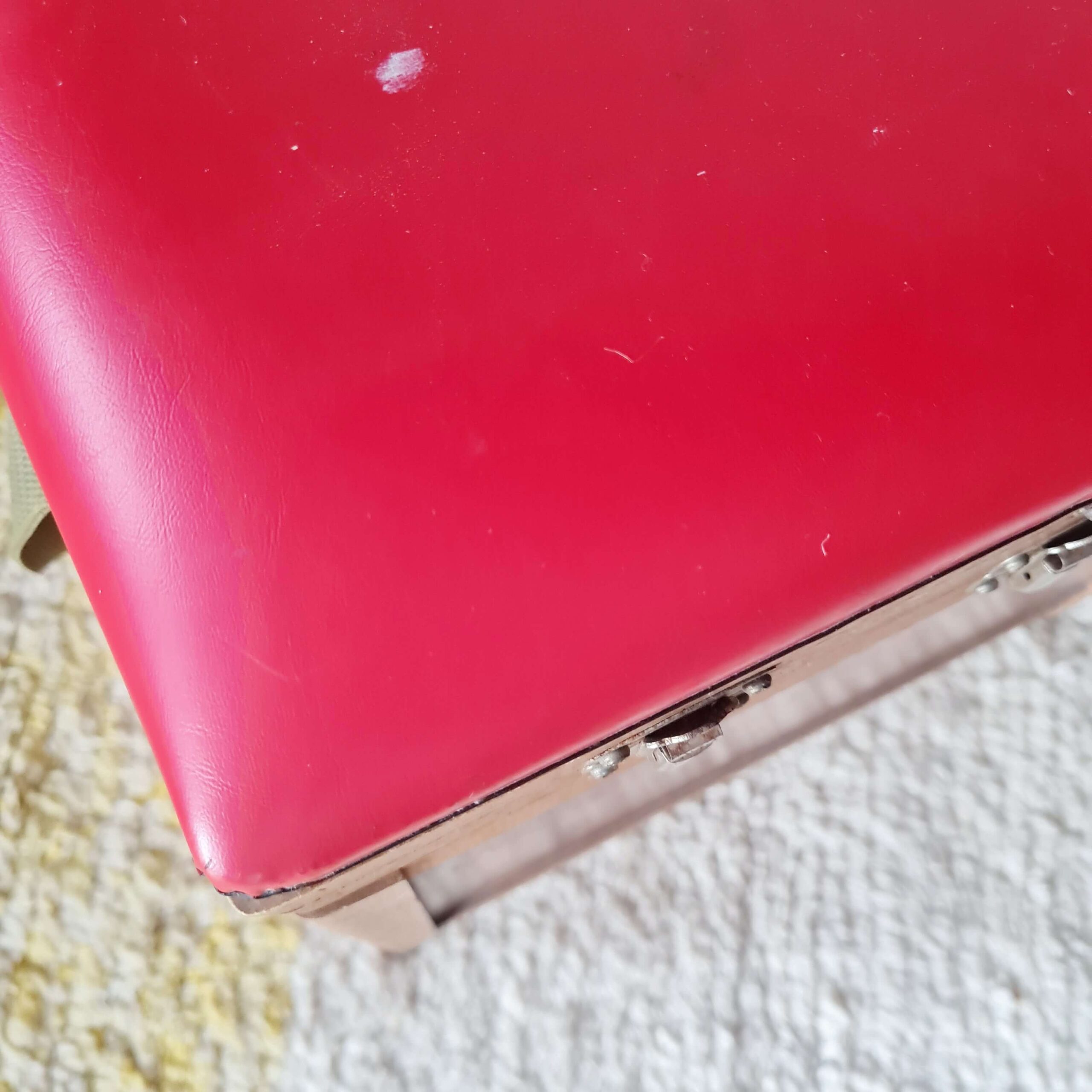 Très gros plan sur un angle de l'assise en simili cuir rouge du tabouret. On voit la jonction avec la structure en bois et un petit éclat sur la peinture blanche de l'arrière-plan.