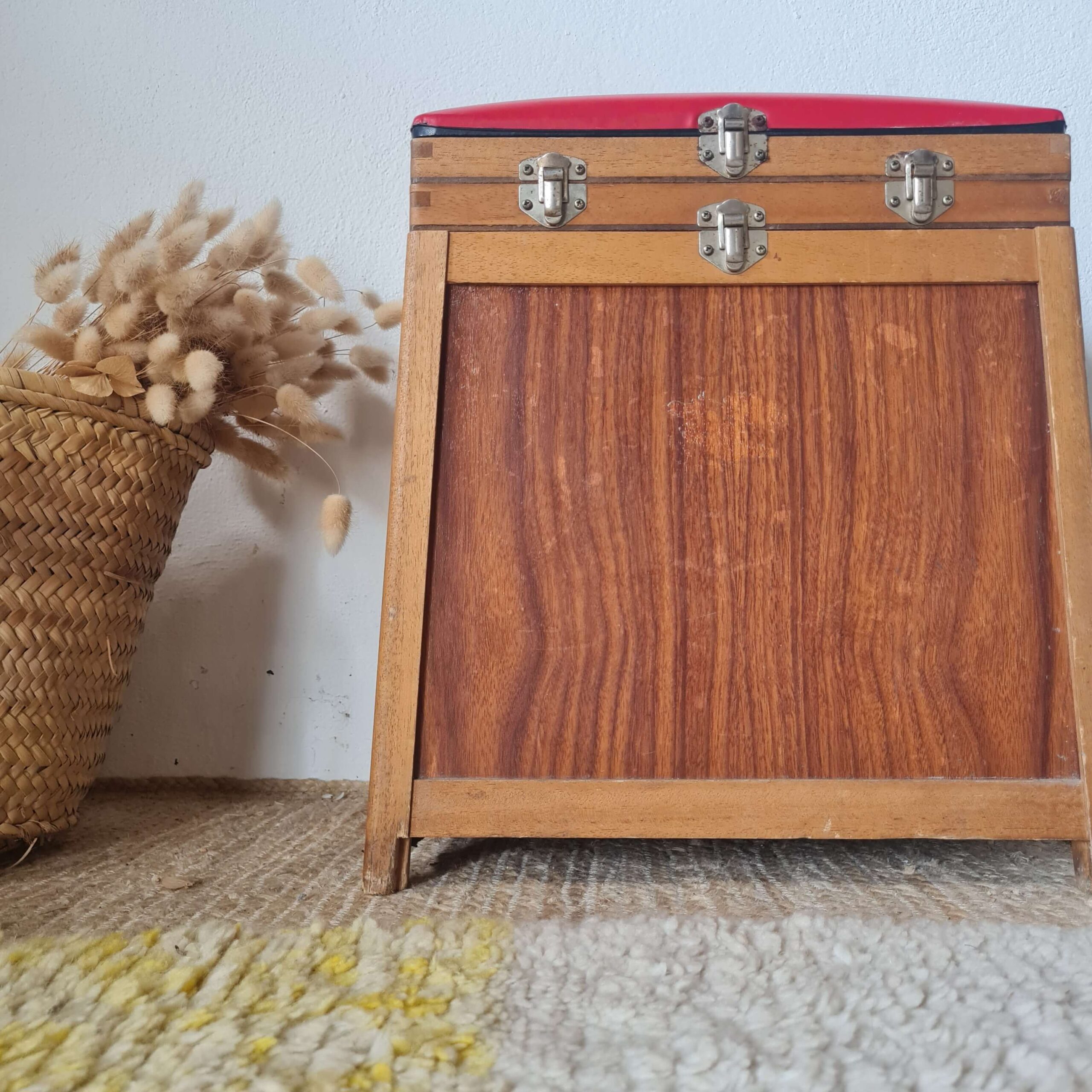 Gros plan sur la façade de l'ancien tabouret de pêcheur. On distingue le veinage du bois, les charnières et les loquets métalliques argentés de l'assise rouge.