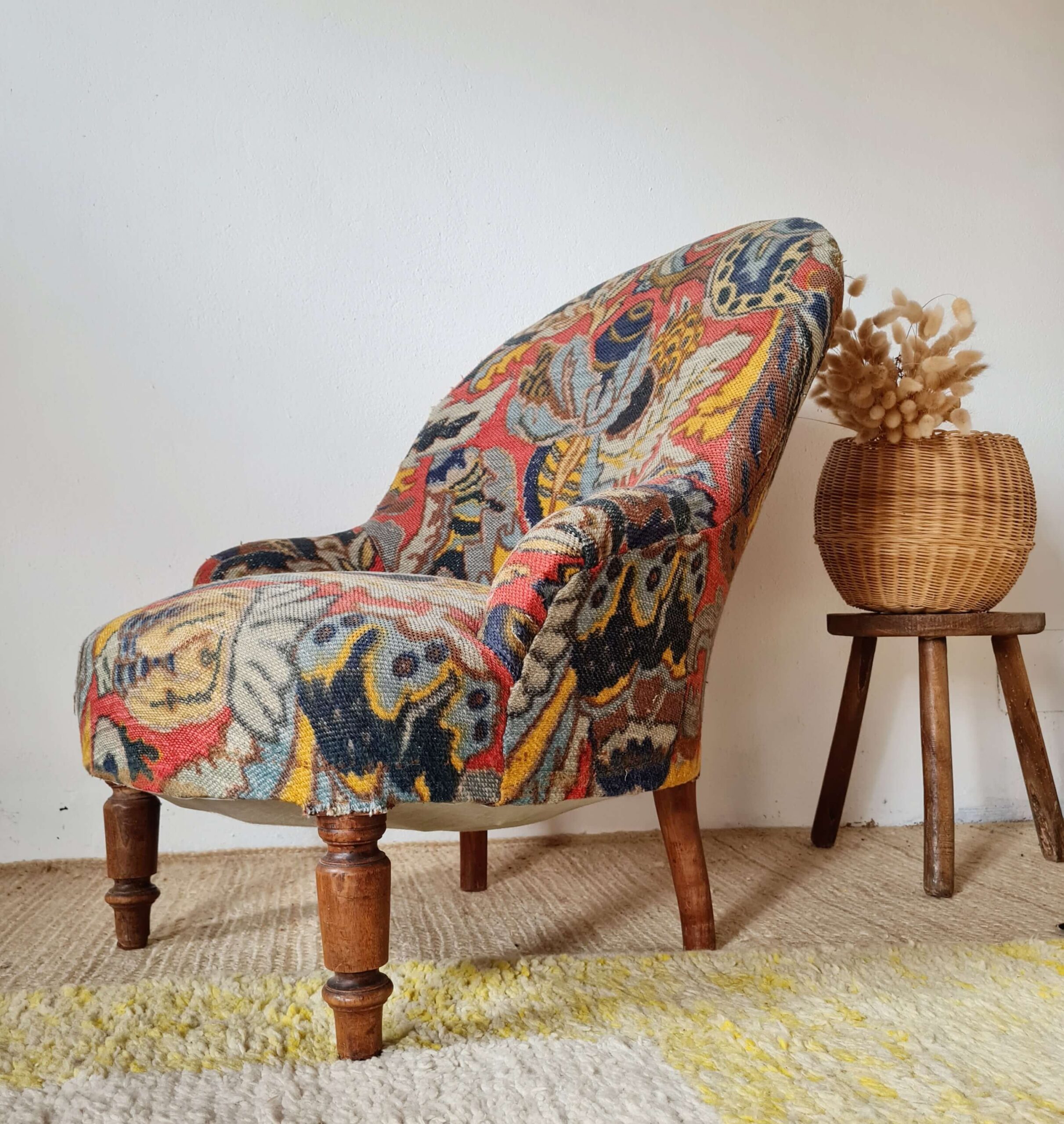 Fauteuil crapaud ancien habillé d'un tissu à motif fleuri logé à côté d'un tabouret tripode ou se tient un vase en rotin vintage